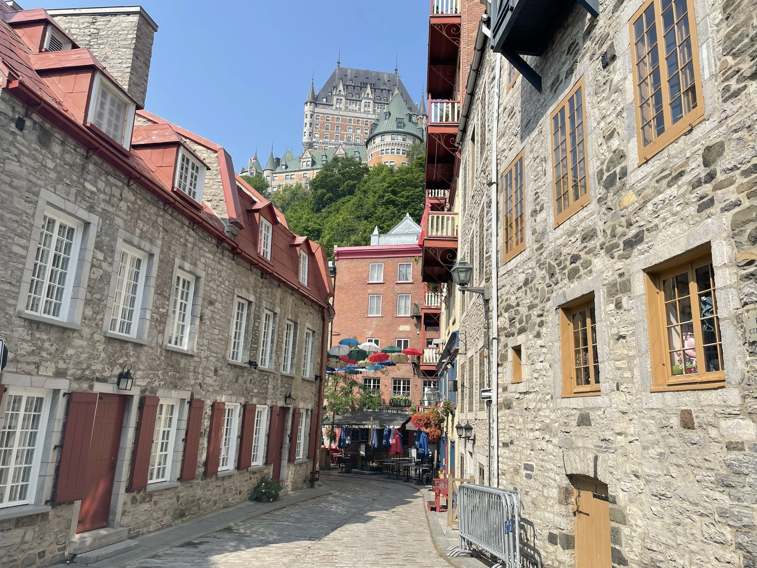 A narrow cobblestone street lined with historic stone buildings, some with red shutters and others with large windows, leading up to a large castle on a hill in the background. Colorful umbrellas are hanging overhead, creating a festive atmosphere.