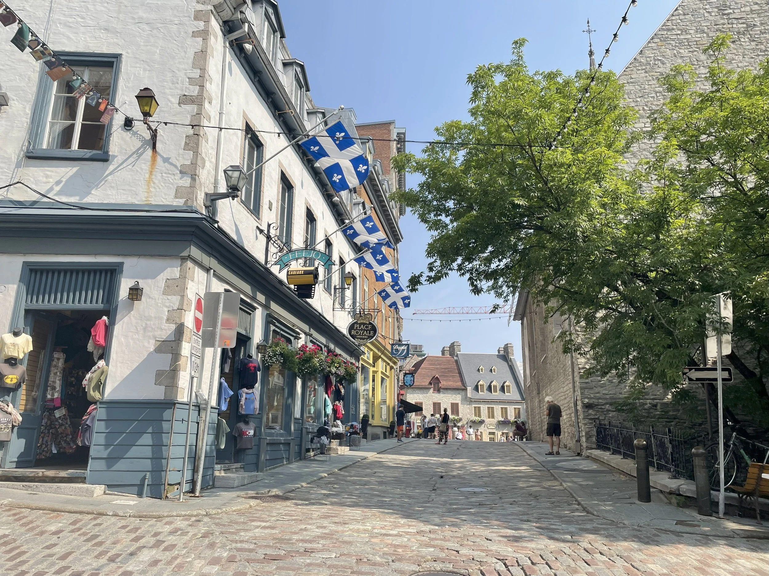 A cobblestone street lined with colorful shops and buildings, with a Canadian flag hanging from a building and people walking along the street on a sunny day with blue skies and green trees.