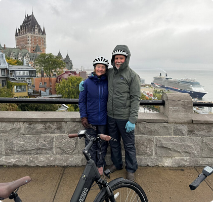 A couple wearing helmets and rain jackets stands together on a stone bridge or walkway, smiling with a cityscape, including a castle and a docked cruise ship, in the background.