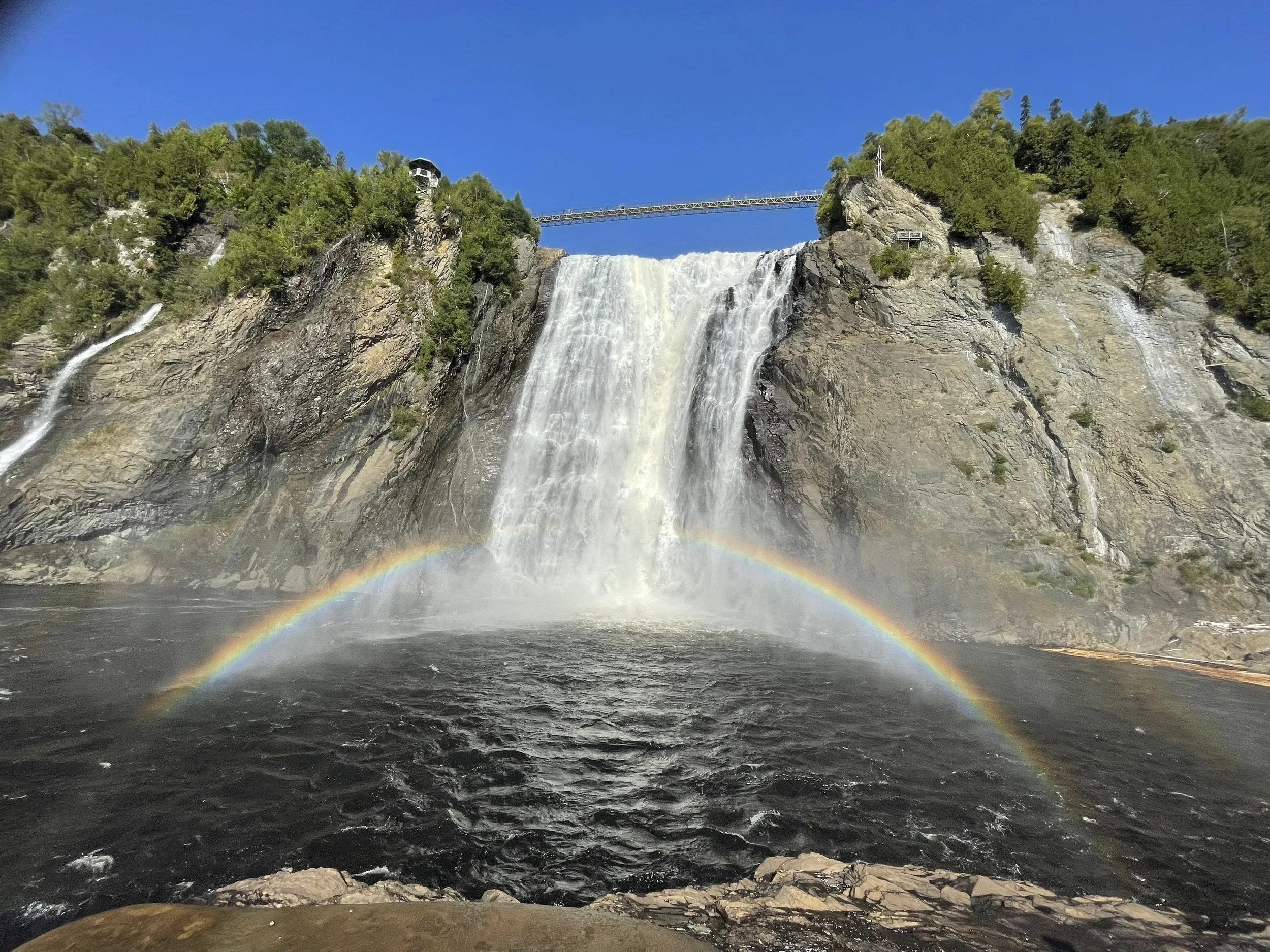 A waterfall with a rainbow at the base, surrounded by rocks and green trees, under a clear blue sky, with a bridge and a viewing platform above.