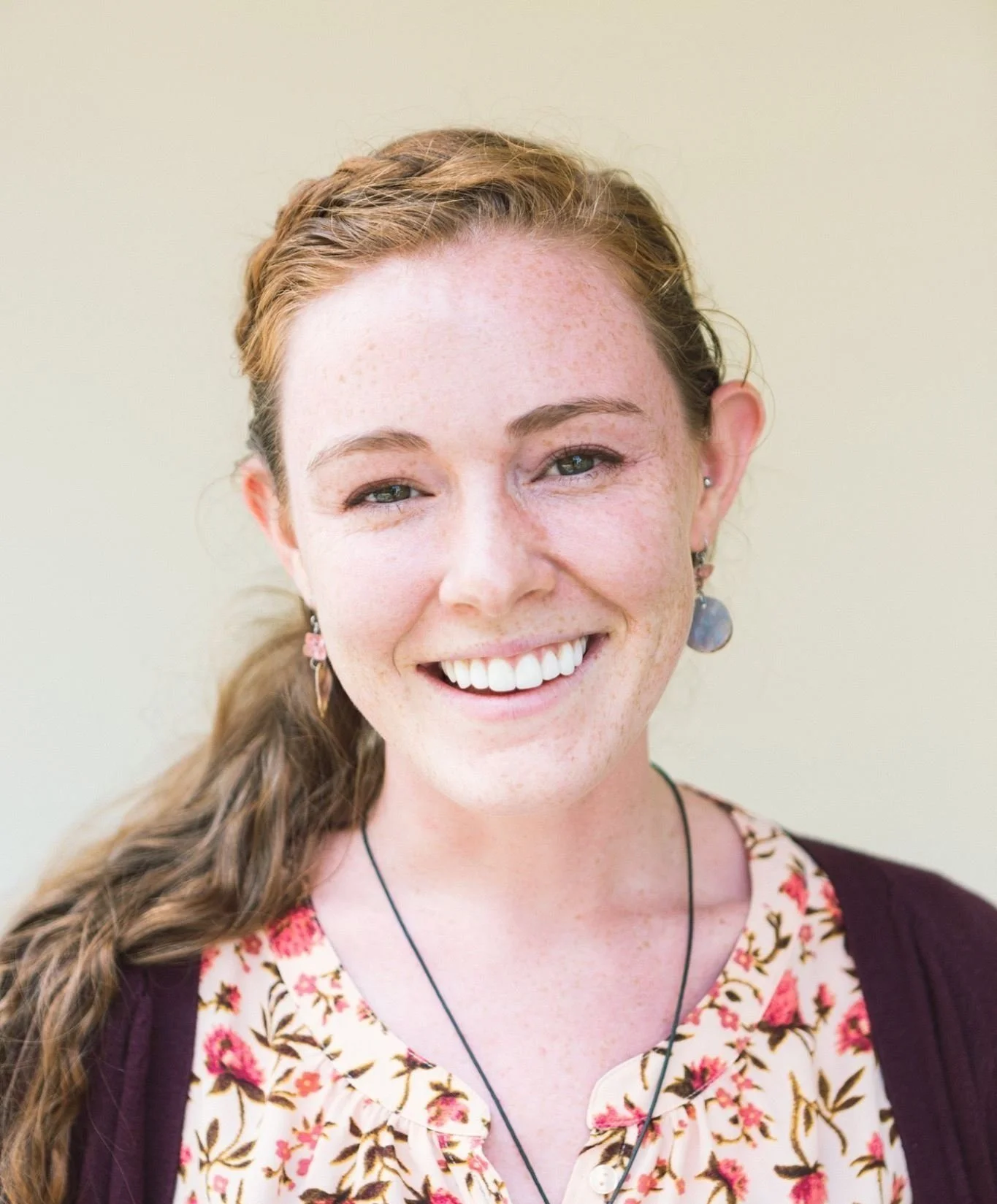 Close-up portrait of a smiling young woman with red hair, freckles, wearing earrings and a floral blouse, against a light background.