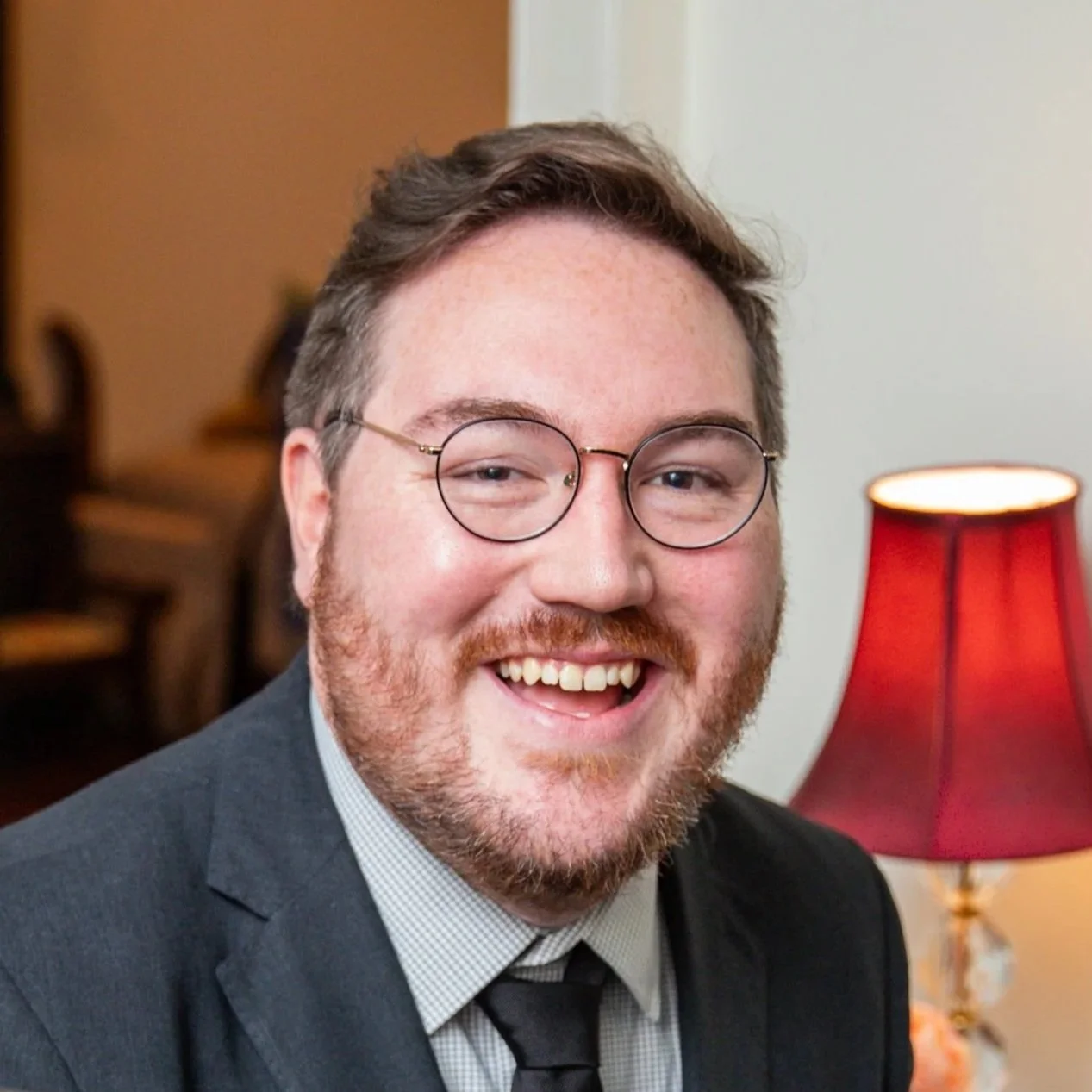 A smiling man with glasses, a beard, and a suit, sitting near a red lamp in an indoor setting.