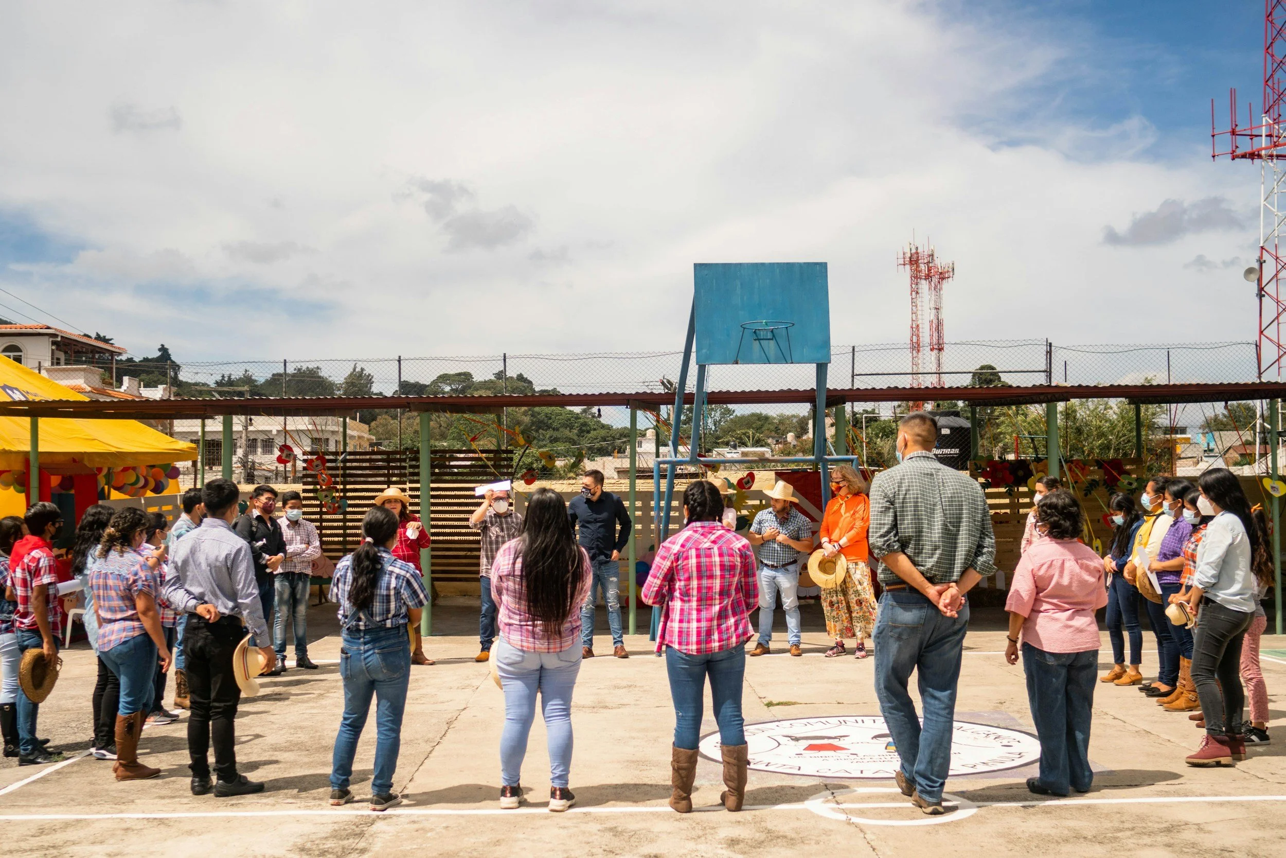 A group of people gathered outdoors in a circle, some taking photos. A blue basketball backboard is visible, and there are festive decorations, with a sunny sky and clouds overhead.