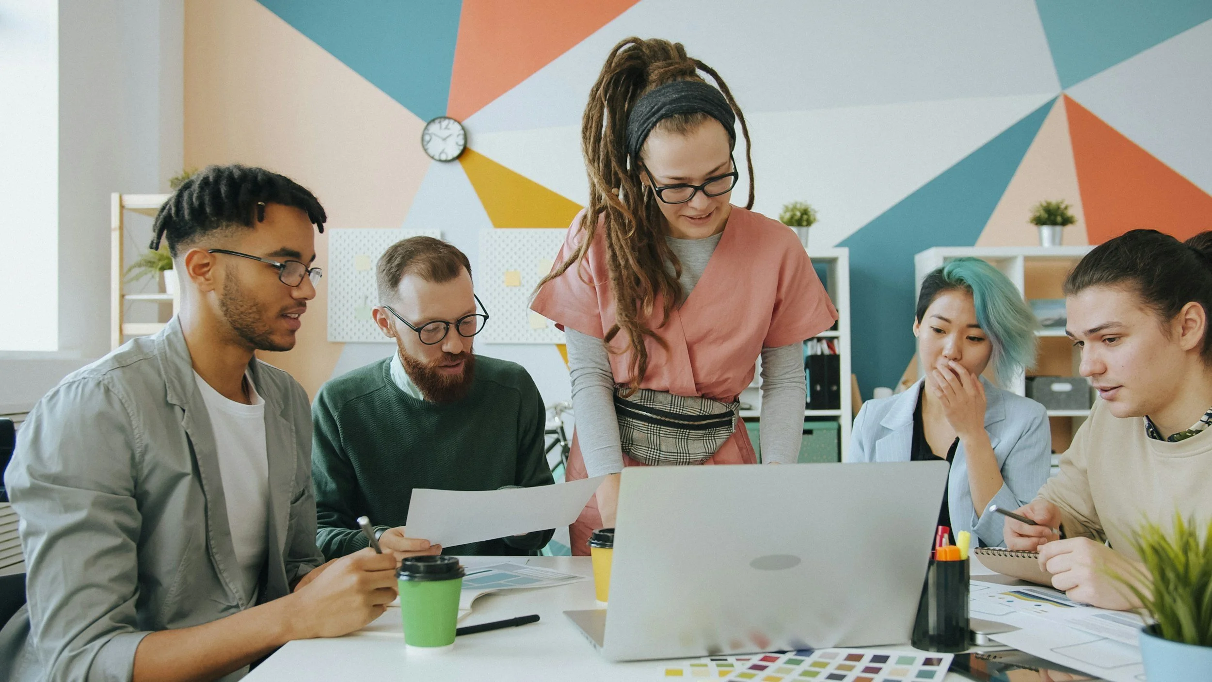 A group discusses a project with computer, papers, tea. Background is colorful and artistic.