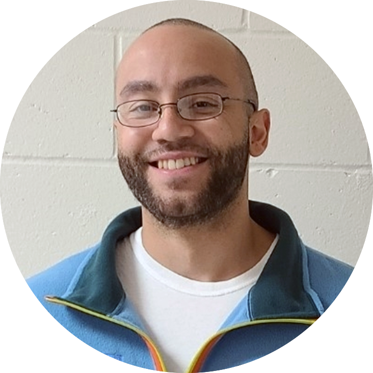 Man with short beard and glasses wearing a blue sweatshirt smiling against a white wall