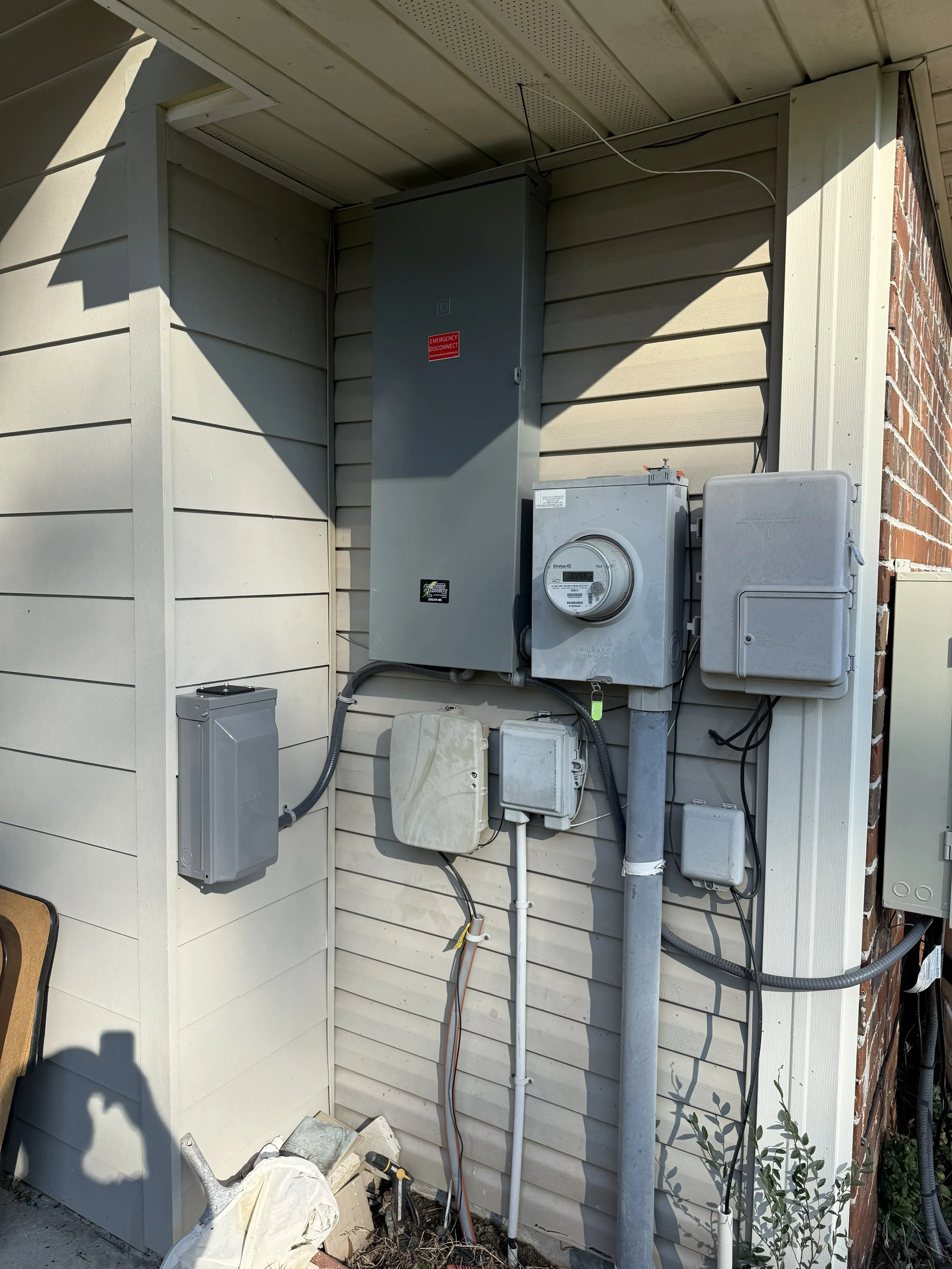 Electrical meter and utility boxes mounted outside a house, with electrical wires and conduits.