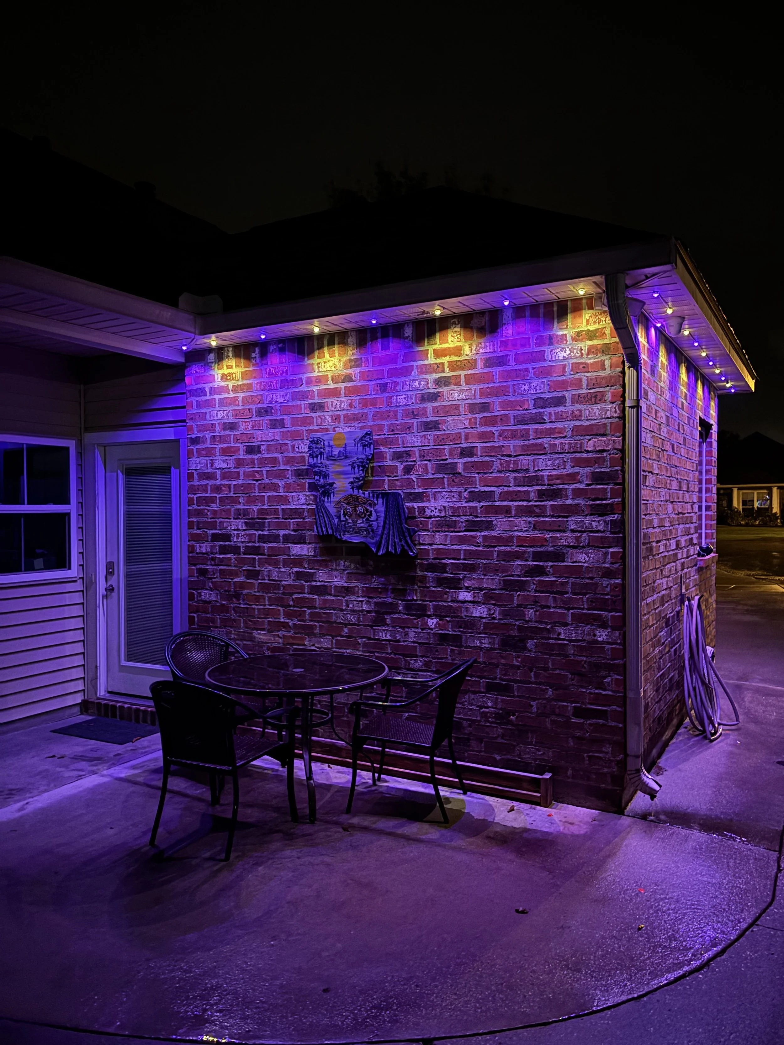 A brick house exterior at night with purple and yellow LED lights on the roof edge. There is a patio table with three chairs on a wet concrete surface. A painting of a cityscape with a sunset hangs on the brick wall.