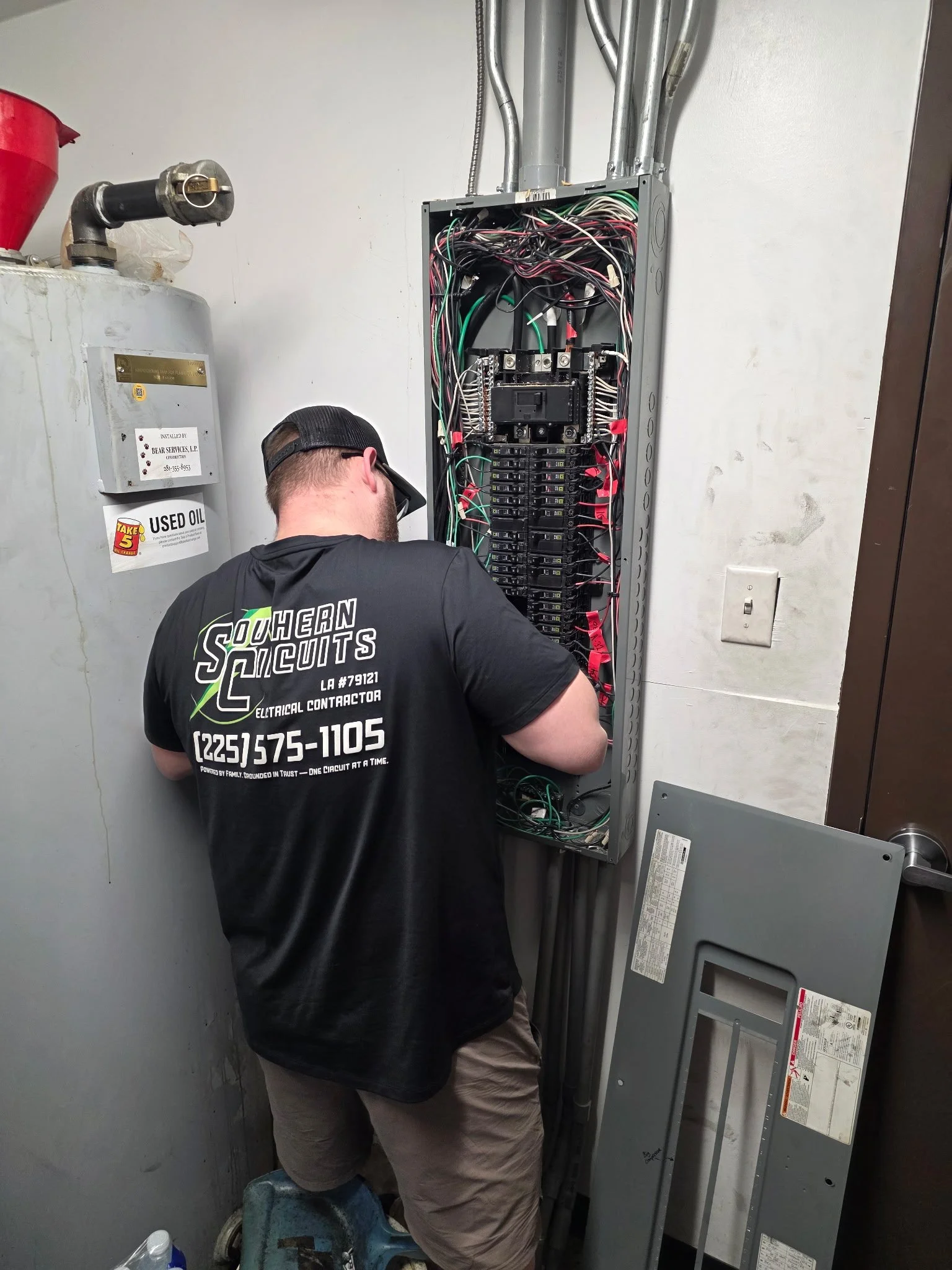 An electrician working on an open electrical panel with wires in a utility room.