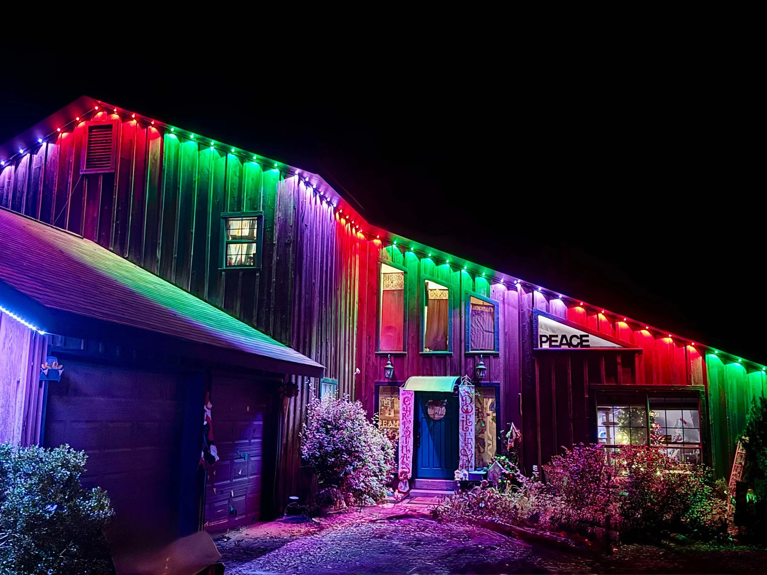 A house decorated with colorful Christmas lights at night, with bushes and a Christmas tree visible in the yard.