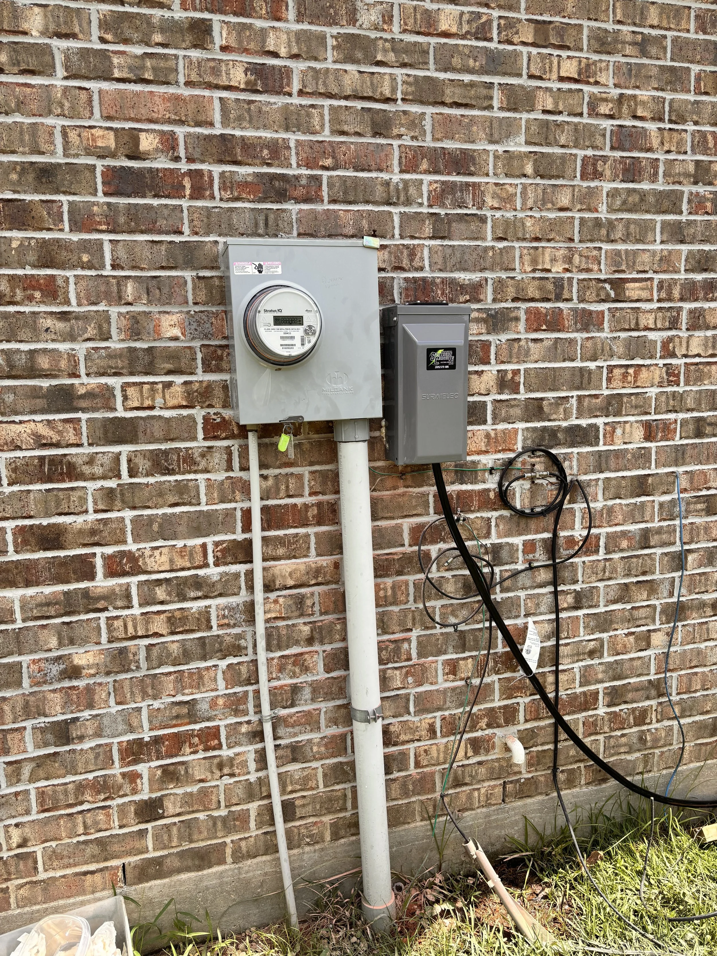 Outdoor electrical meter and electrical box attached to a brick wall, with cables hanging and a small patch of grass at the bottom.