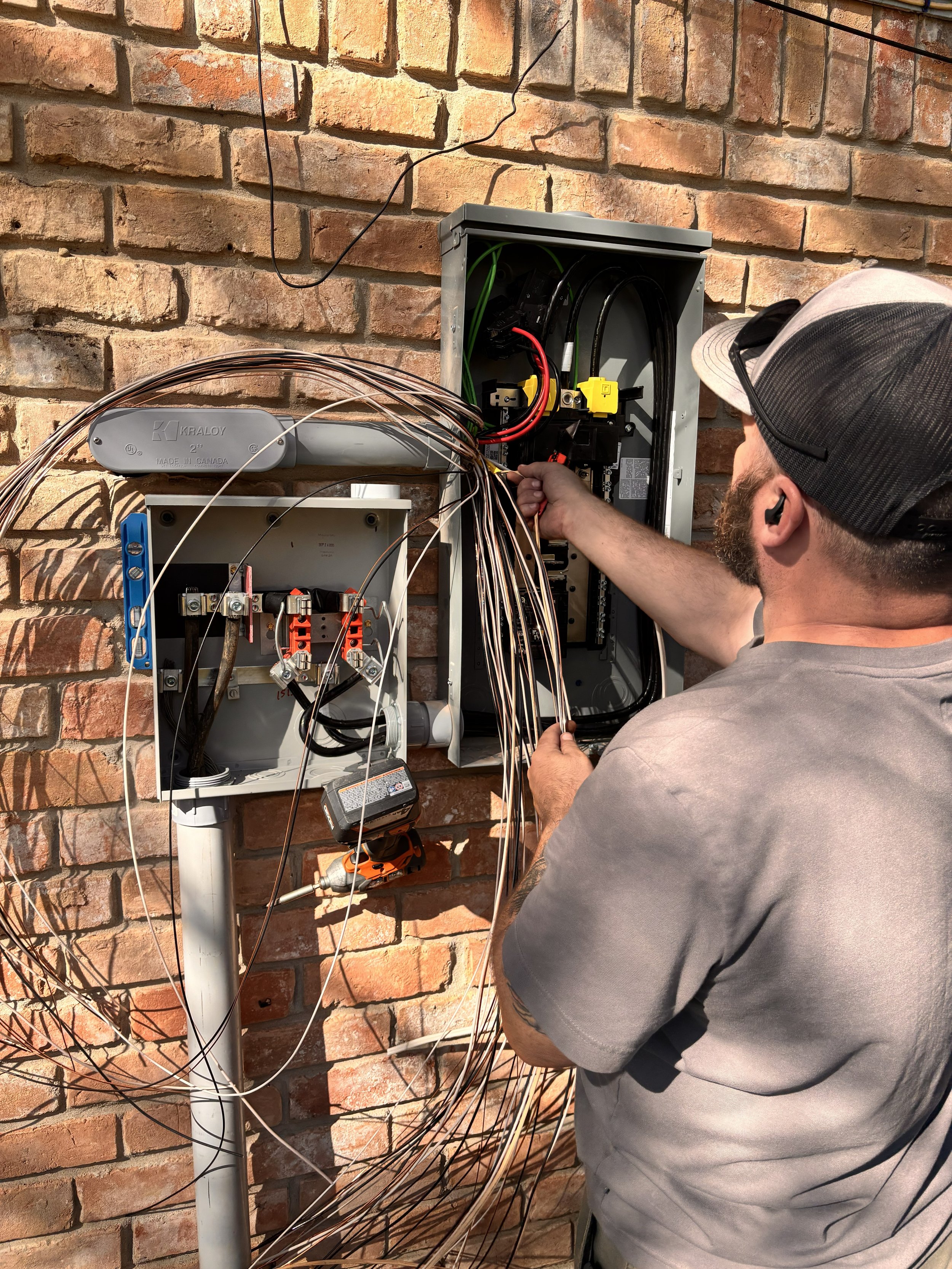 A technician works on an electrical panel mounted on a brick wall, with numerous wires connected to the panel.