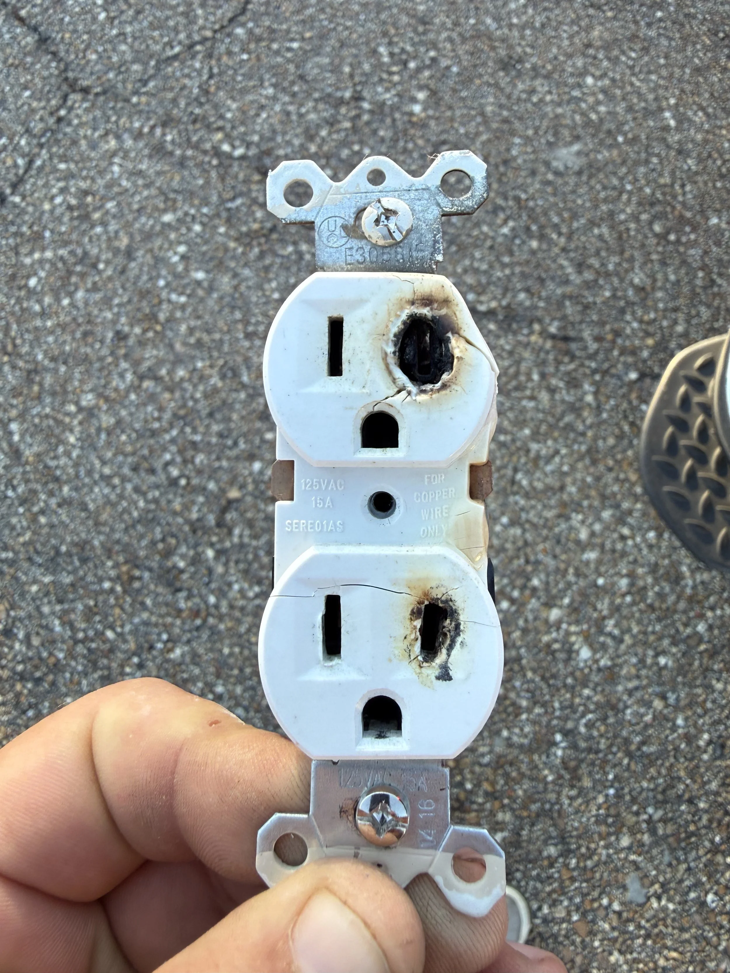 A hand holding a damaged electrical outlet splitter with one side burned and blackened, showing signs of electrical damage.