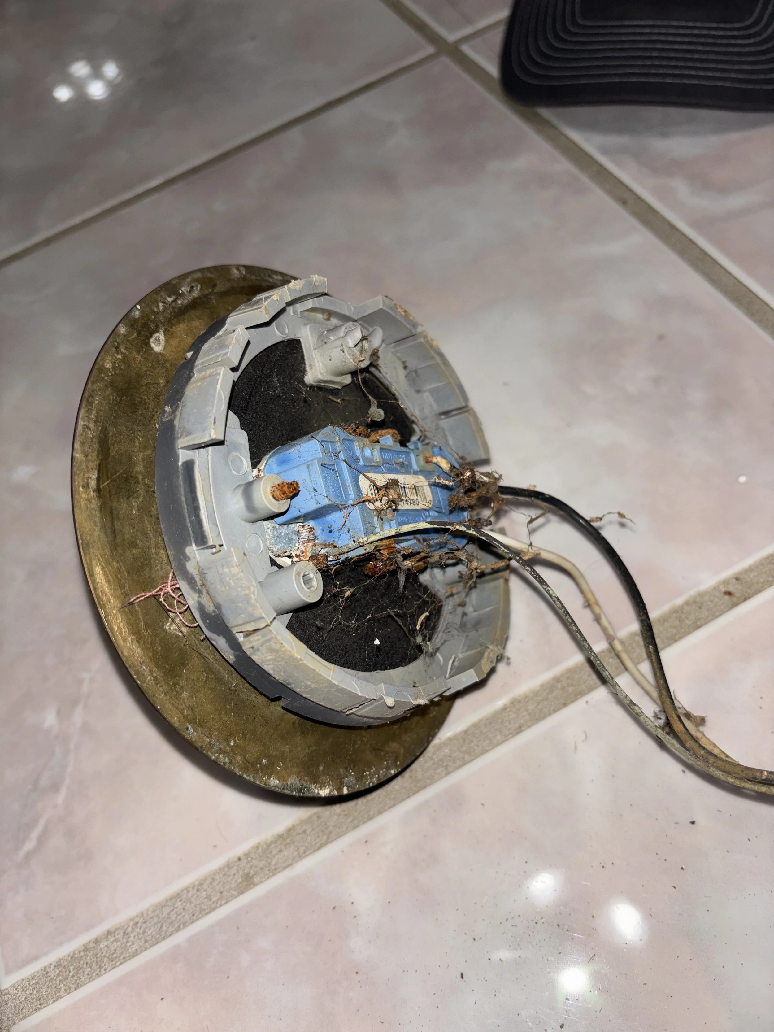 An old, damaged ceiling light fixture with exposed wires and rust, lying on a tiled floor.
