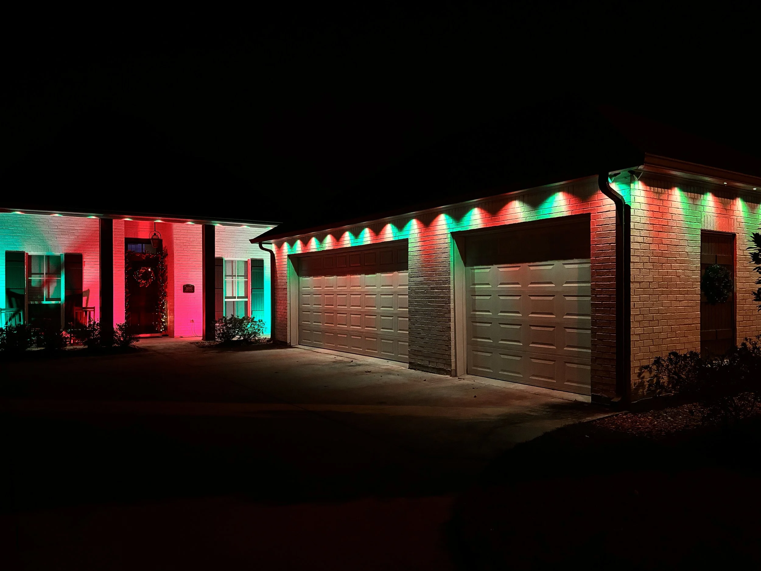 A house decorated with red and green Christmas lights, with the lights outlining the roofline and a wreath hanging on the front door at night.
