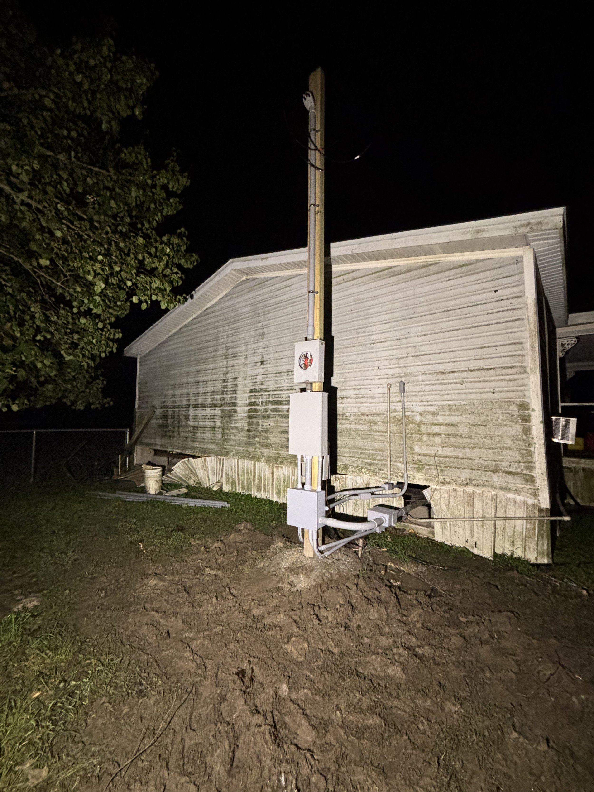 Back of a weathered house with electrical meters and cables, night scene, partially muddy ground.