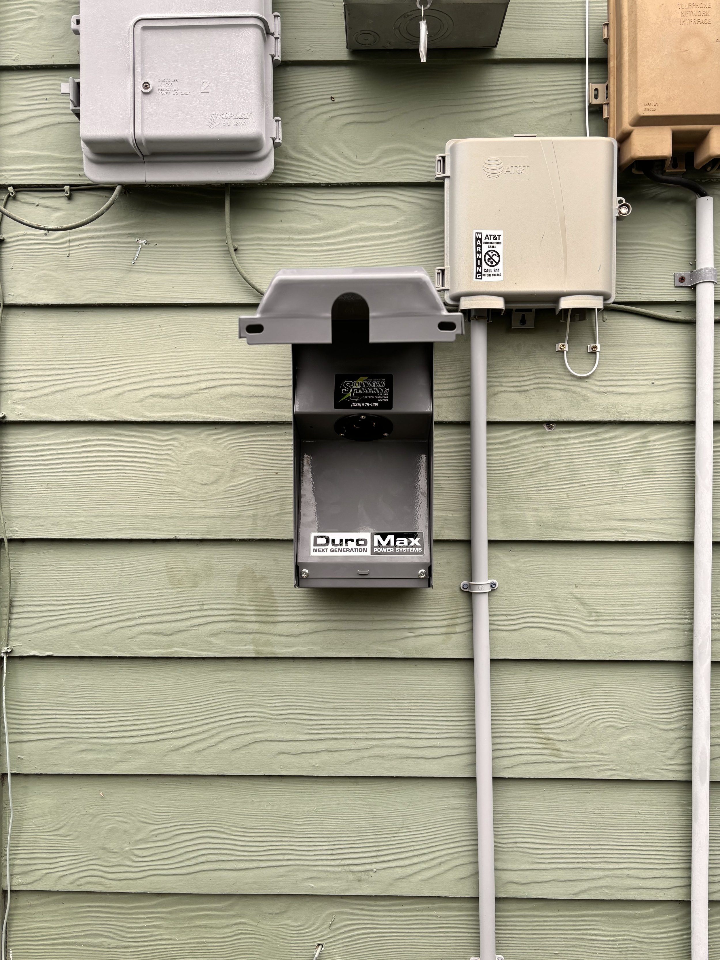 Electrical and communication boxes mounted on a green wooden wall, with visible wires and conduit.