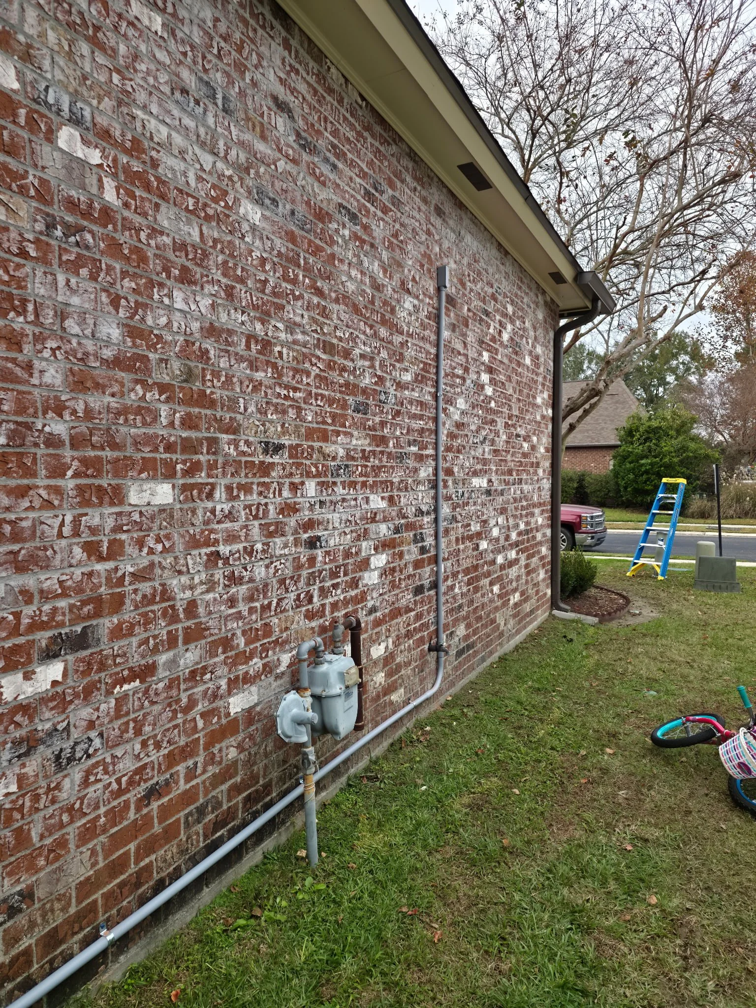 A brick house exterior with gas meter and pipes, a ladder, a parked truck, and children's bicycle in the yard.