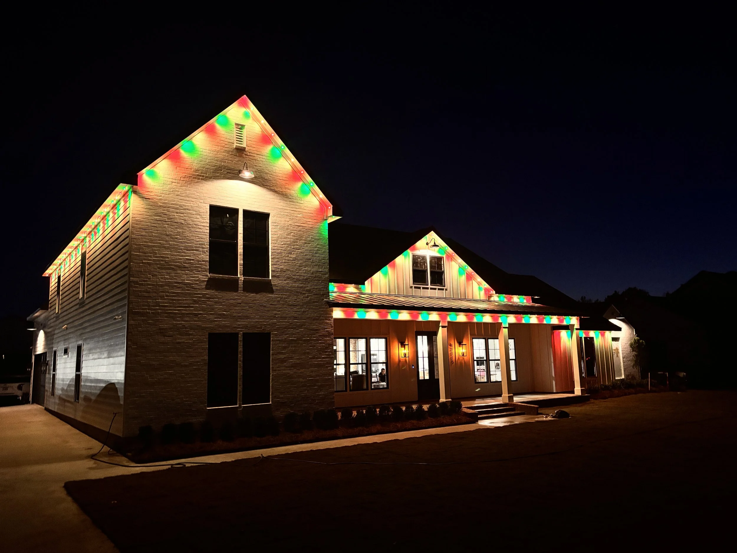 A two-story house decorated with multicolored Christmas lights at night, with a dark sky in the background.