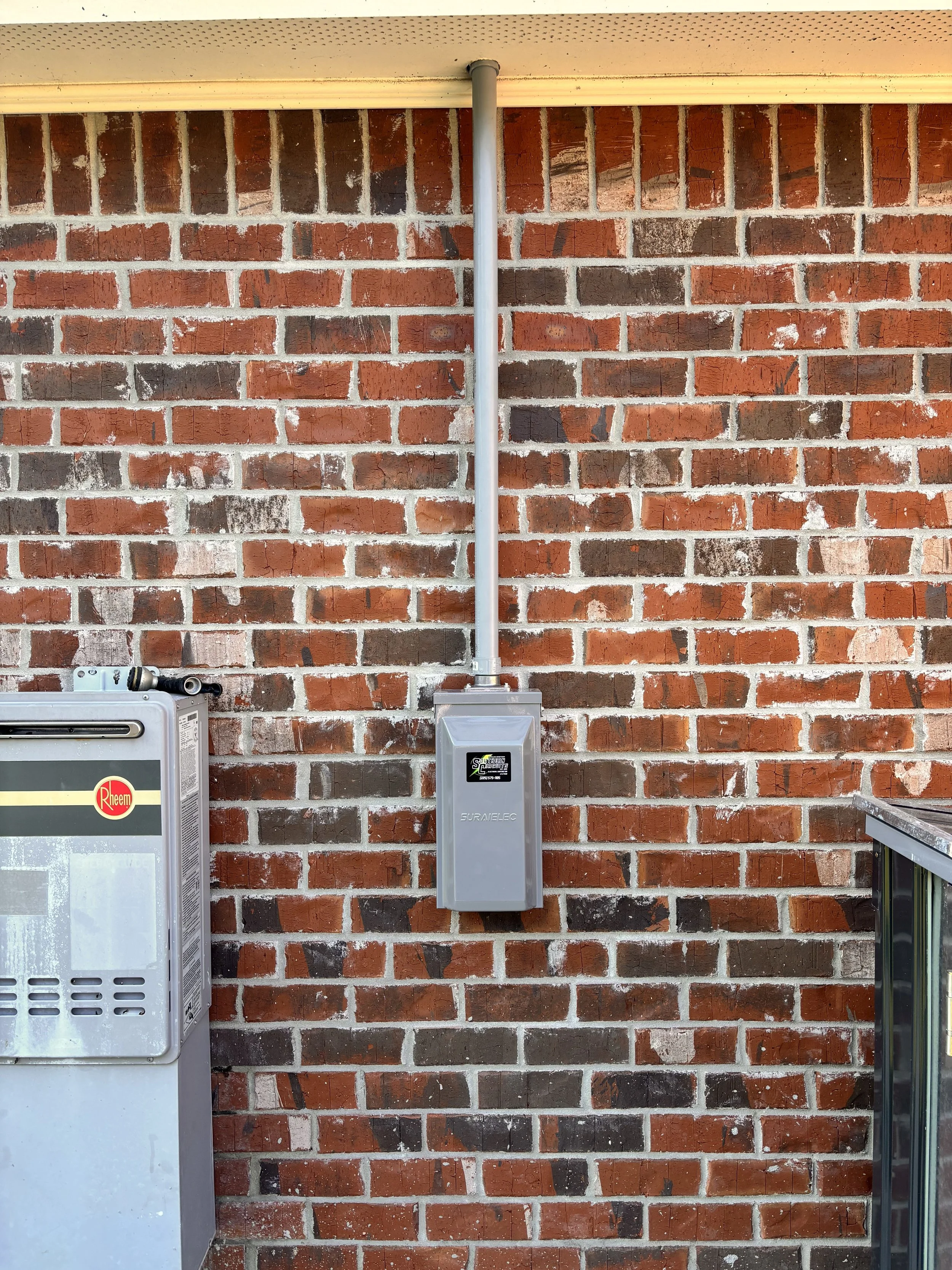 Close-up of a brick wall with an electrical box and conduit pipe mounted on it, with part of a metal utility box visible on the left and a countertop edge on the right.