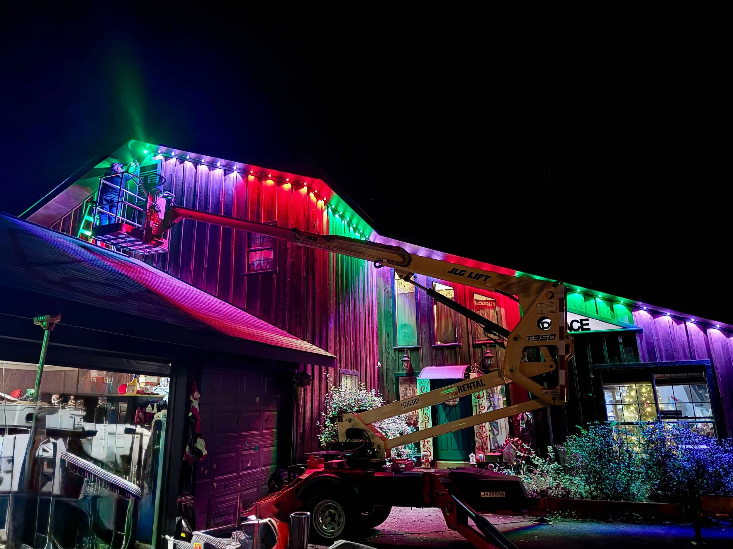 A building decorated with multicolored Christmas lights at night, with a cherry picker lift near the entrance.