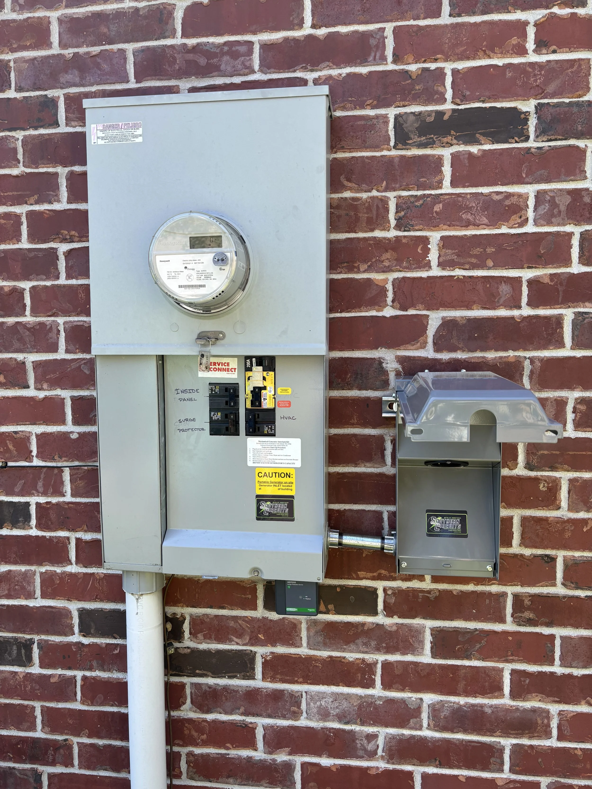 Electric meter and electrical panel mounted on a brick wall with a gray protective enclosure and a small gray utility box nearby.