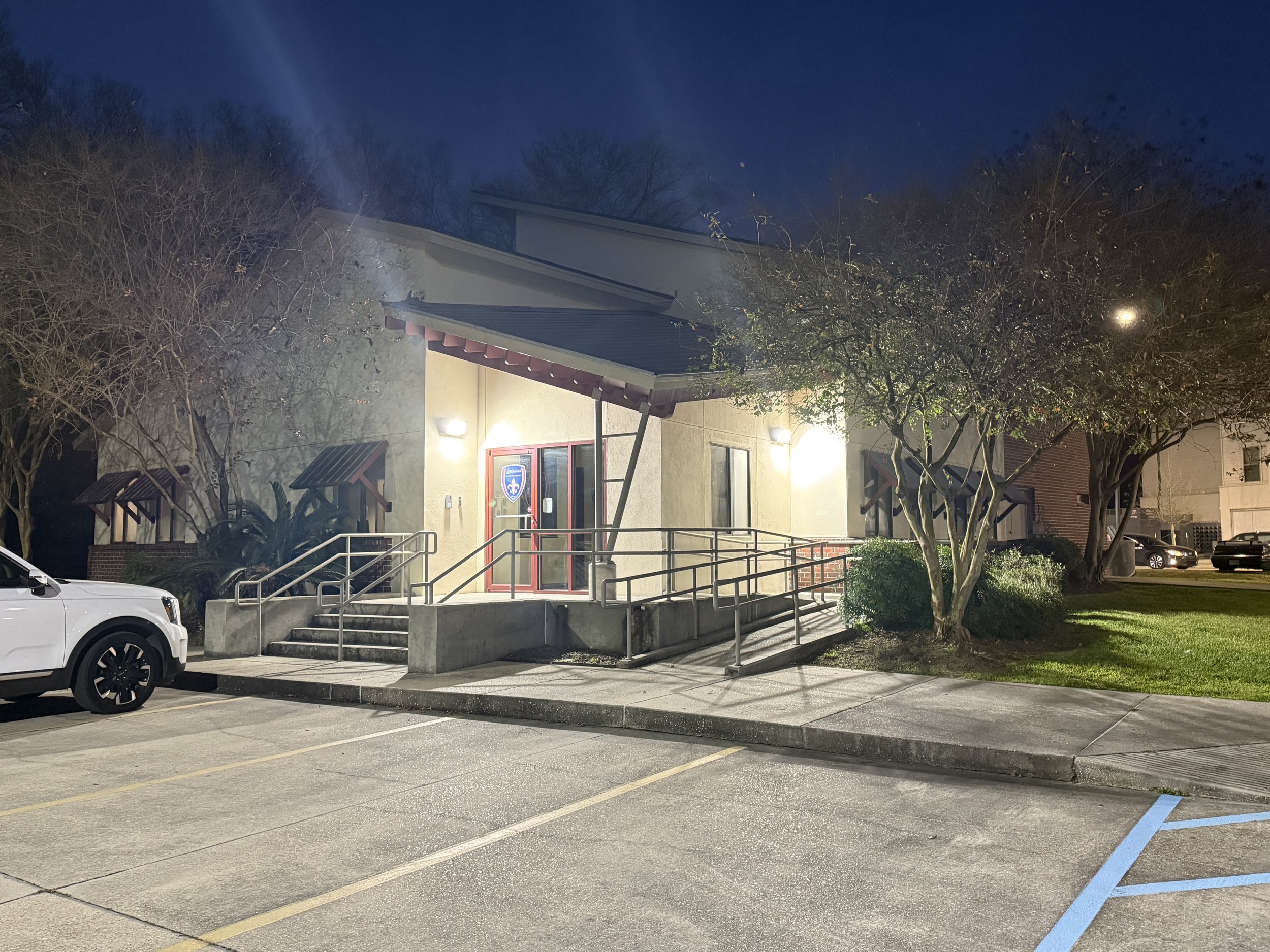 A single-story building with beige walls and a dark roof illuminated at night. It has a front door with a blue sign and steps leading up to the entrance. There are trees and bushes nearby, and a parking lot with a white vehicle parked in front.