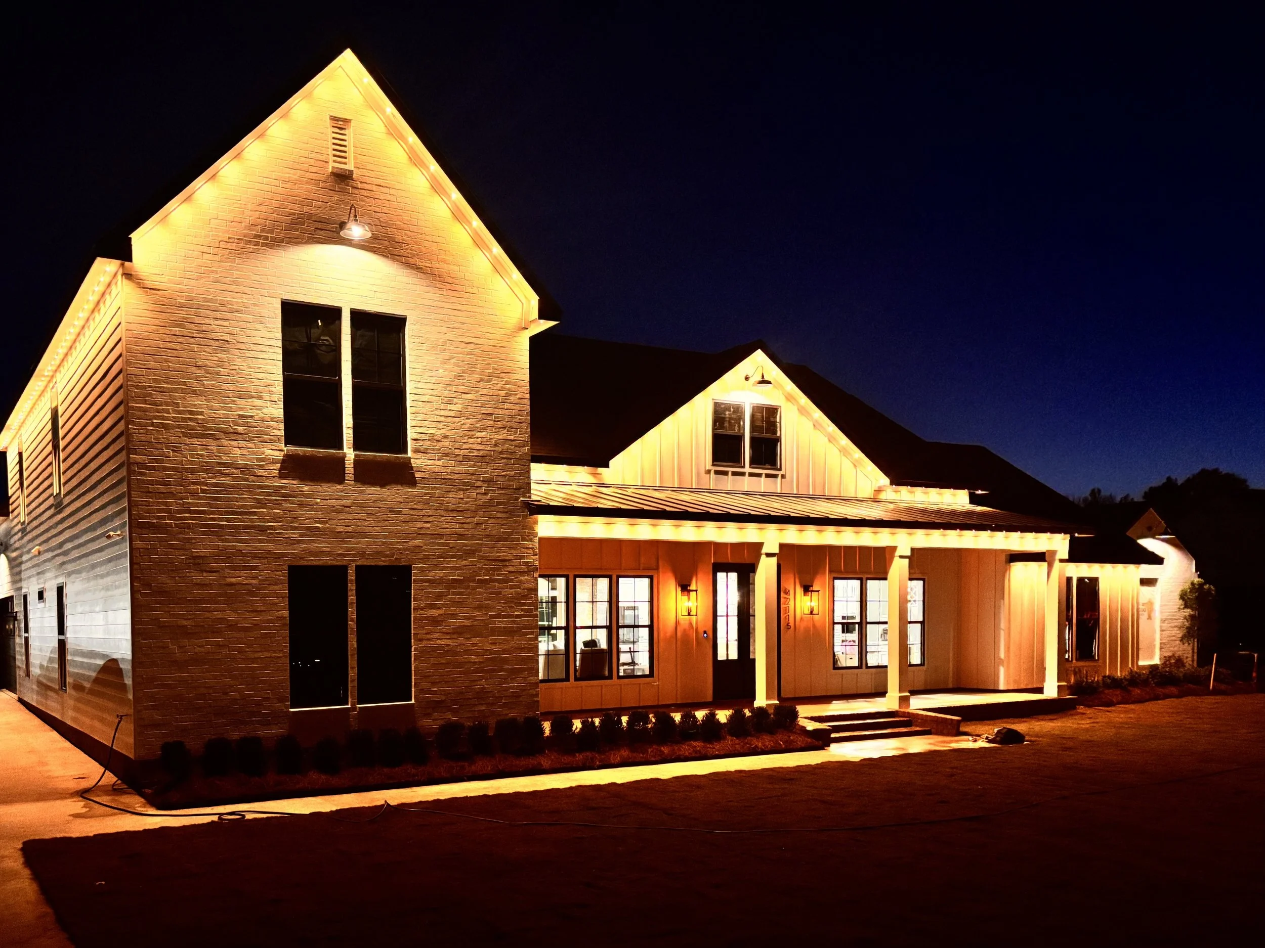 A modern house illuminated with exterior lighting at night, featuring brick and siding facade, multiple windows, front porch, and a well-lit yard.
