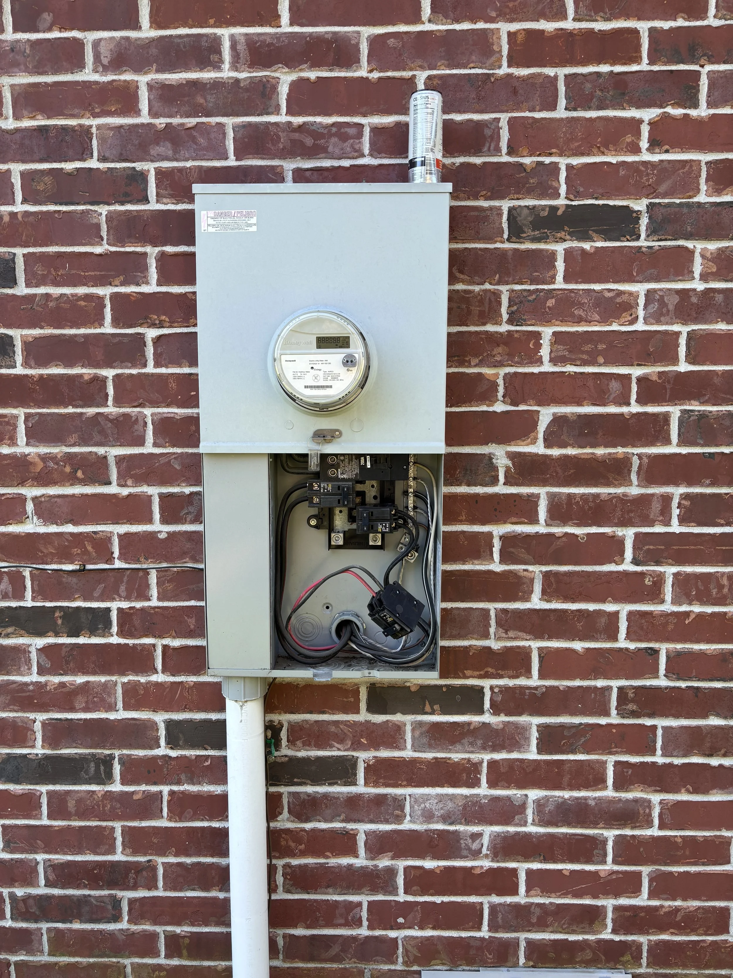 An outdoor electrical meter box mounted on a brick wall with its cover open, revealing wiring and components inside, and a white pipe extending downwards from the bottom.