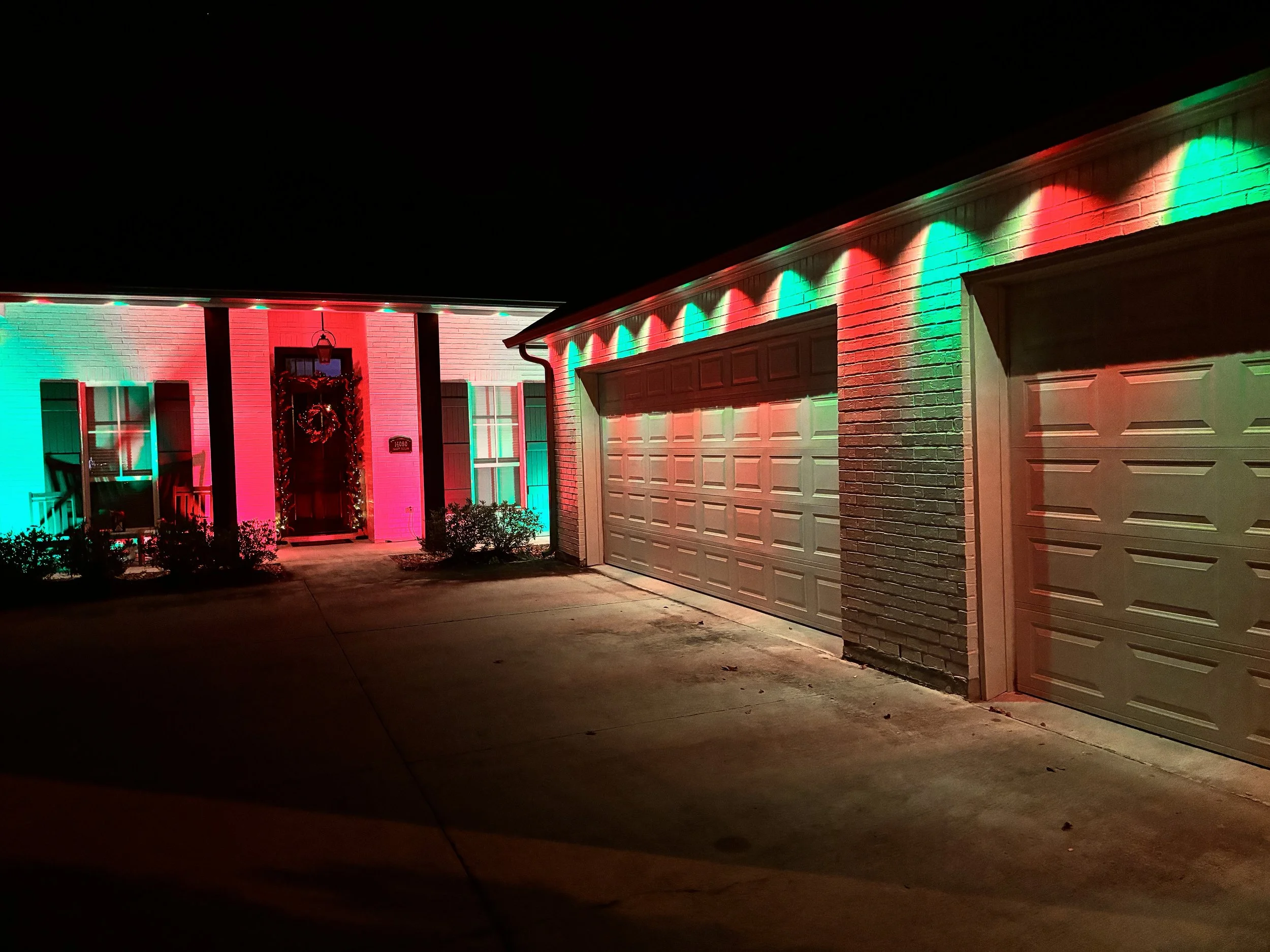 Nighttime view of a house decorated with red and green Christmas lights, with the garage and front door illuminated by colorful lights, and a wreath hanging on the front door.