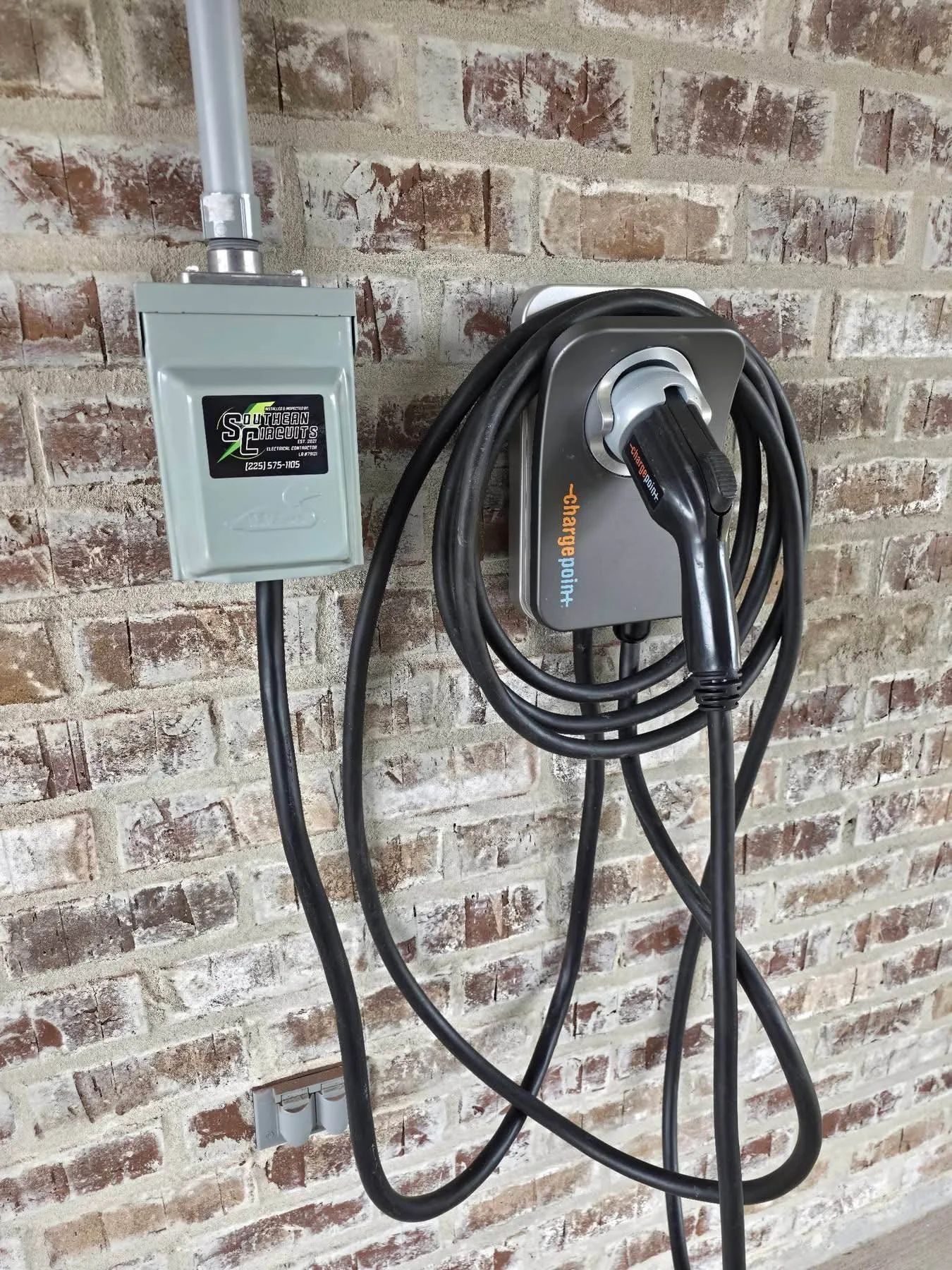 Electric vehicle charging station connected to a gray electrical box, mounted on a brick wall.