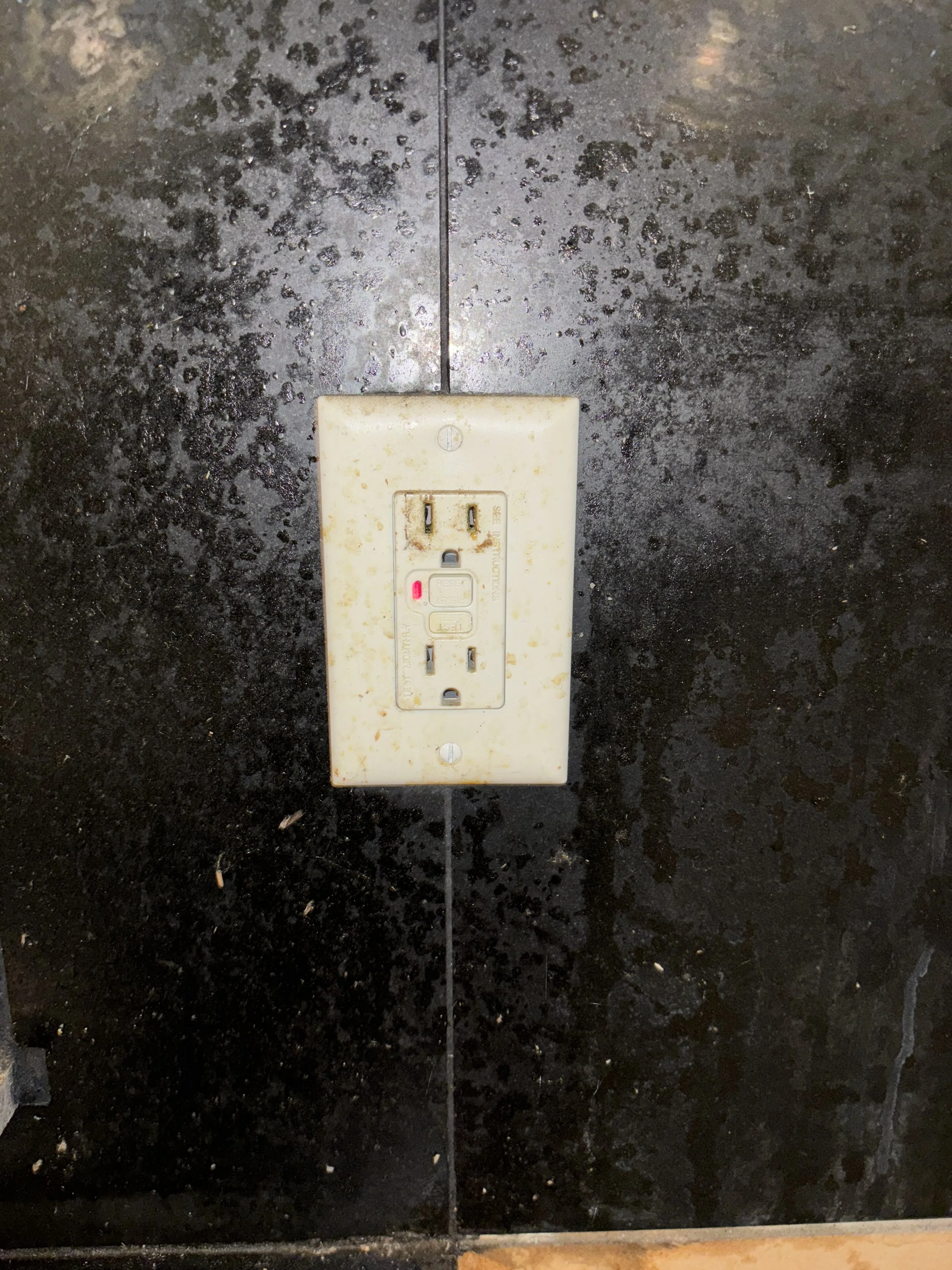 A close-up of a dirty electrical outlet on a black tiled wall with some debris around it.