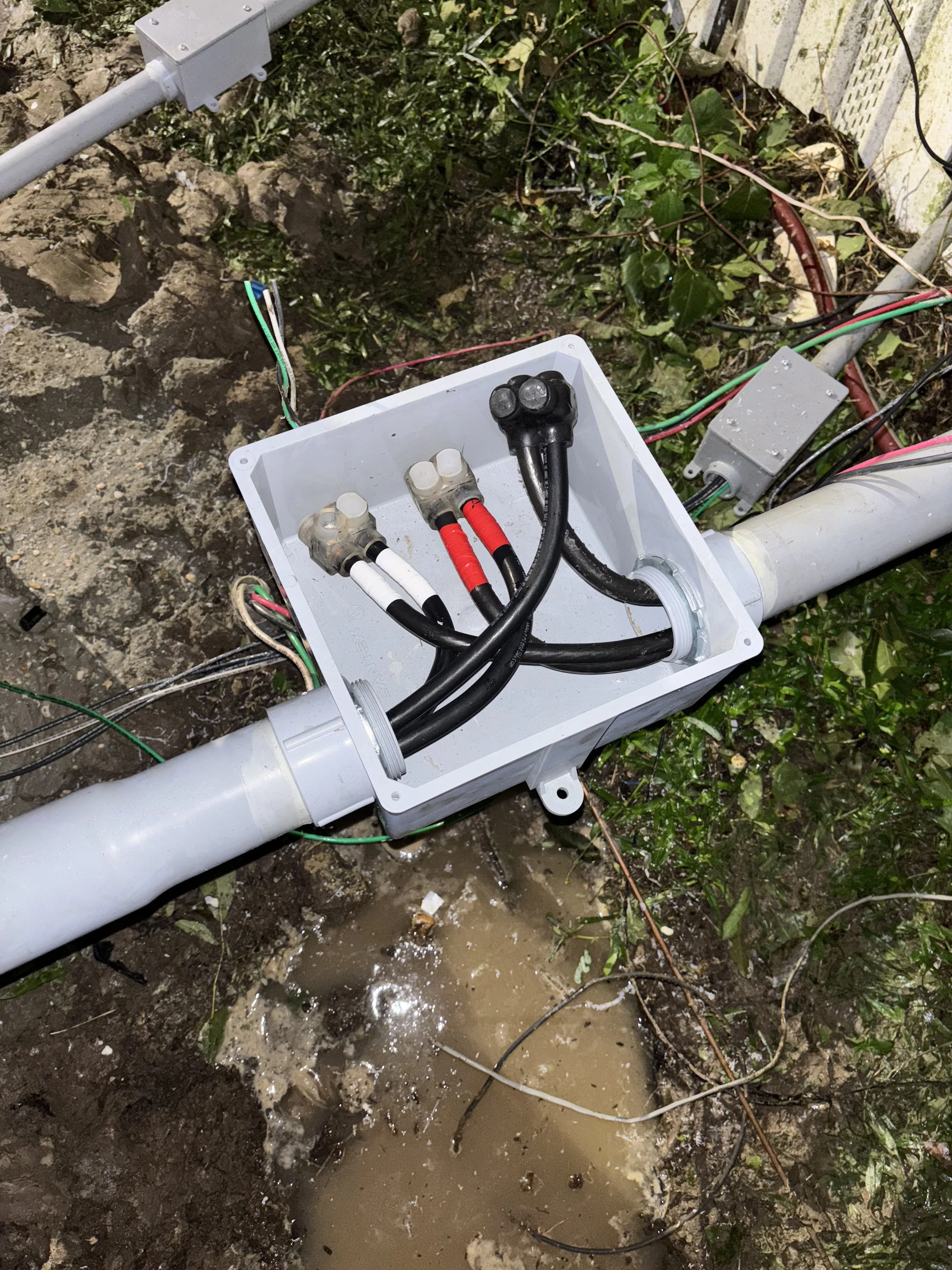 Close-up of an outdoor electrical junction box with electrical cables and wiring, mounted on a pipe underground near muddy ground and grass.