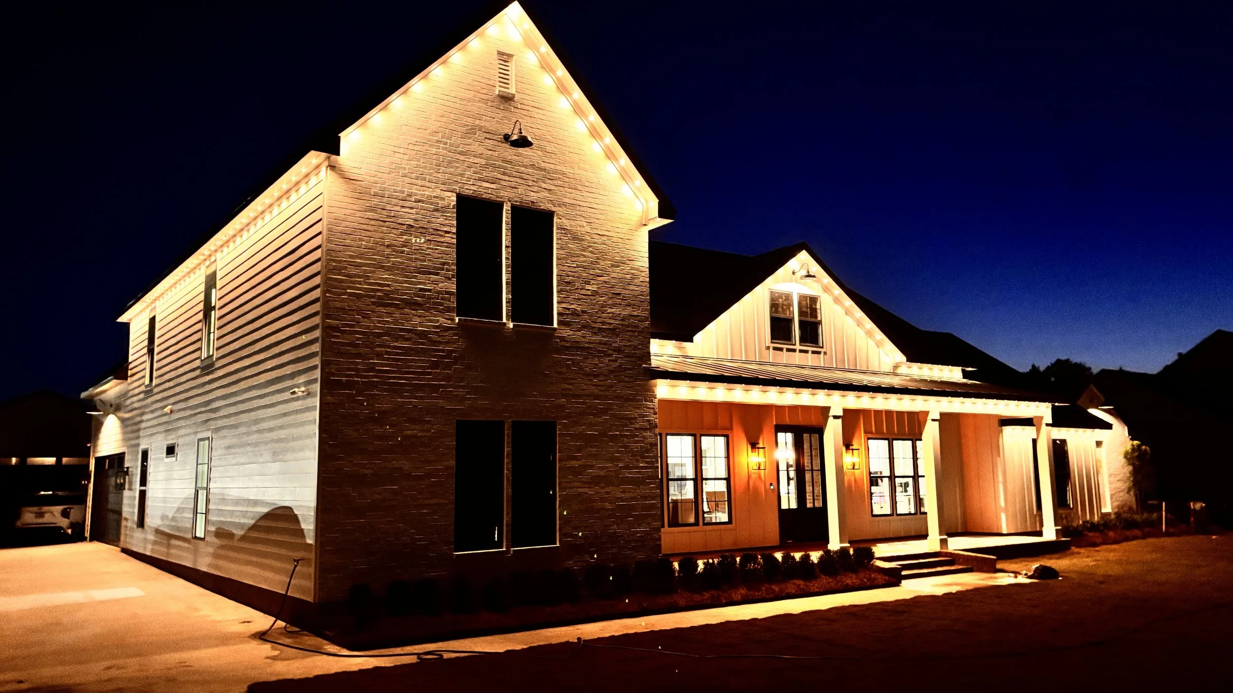 Illuminated two-story house at night with string lights along the roofline, brick and white siding exterior, front porch with steps, and multiple windows.