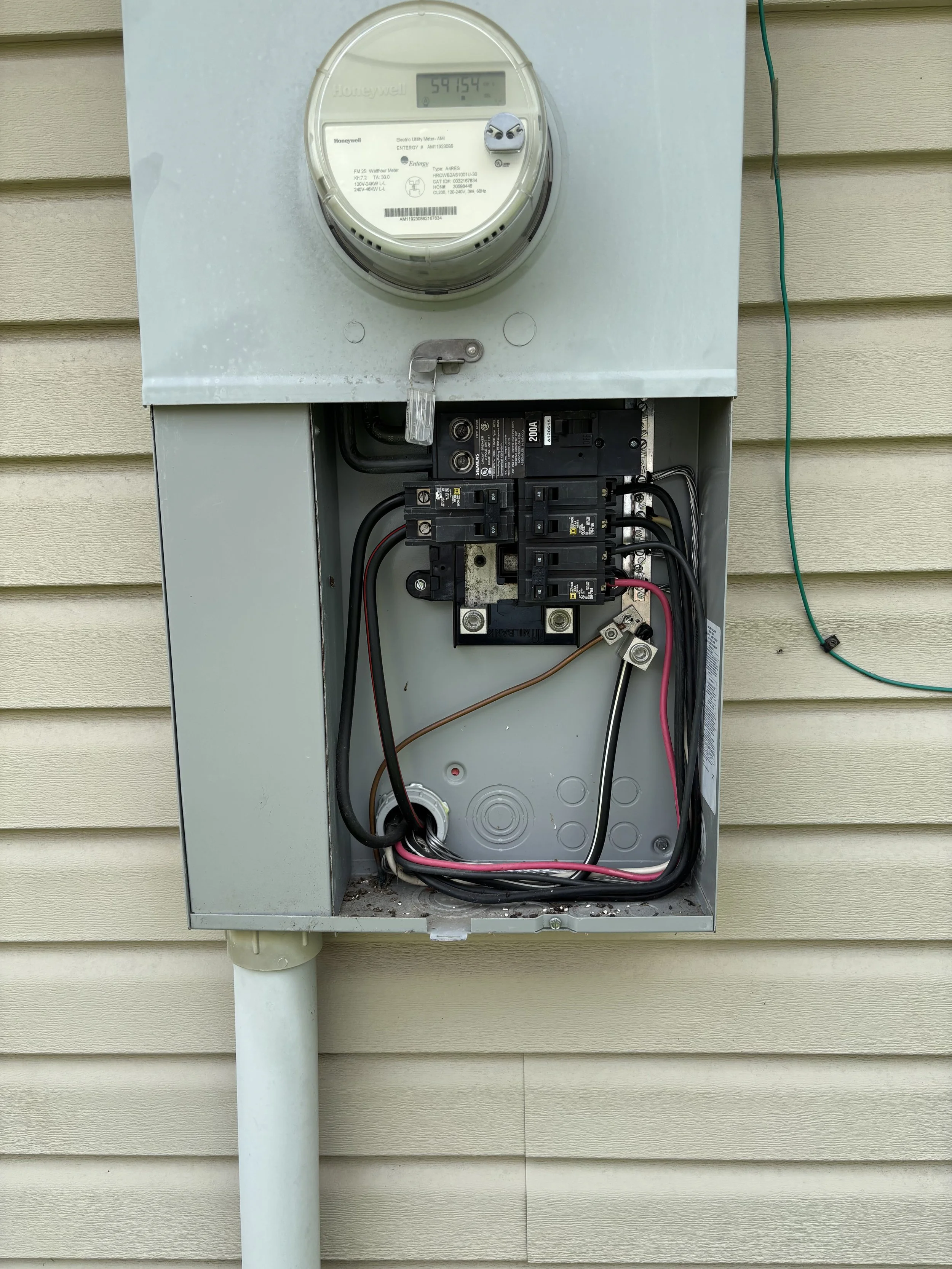 Open electric meter box with wiring and circuit breakers, outside house with beige siding.