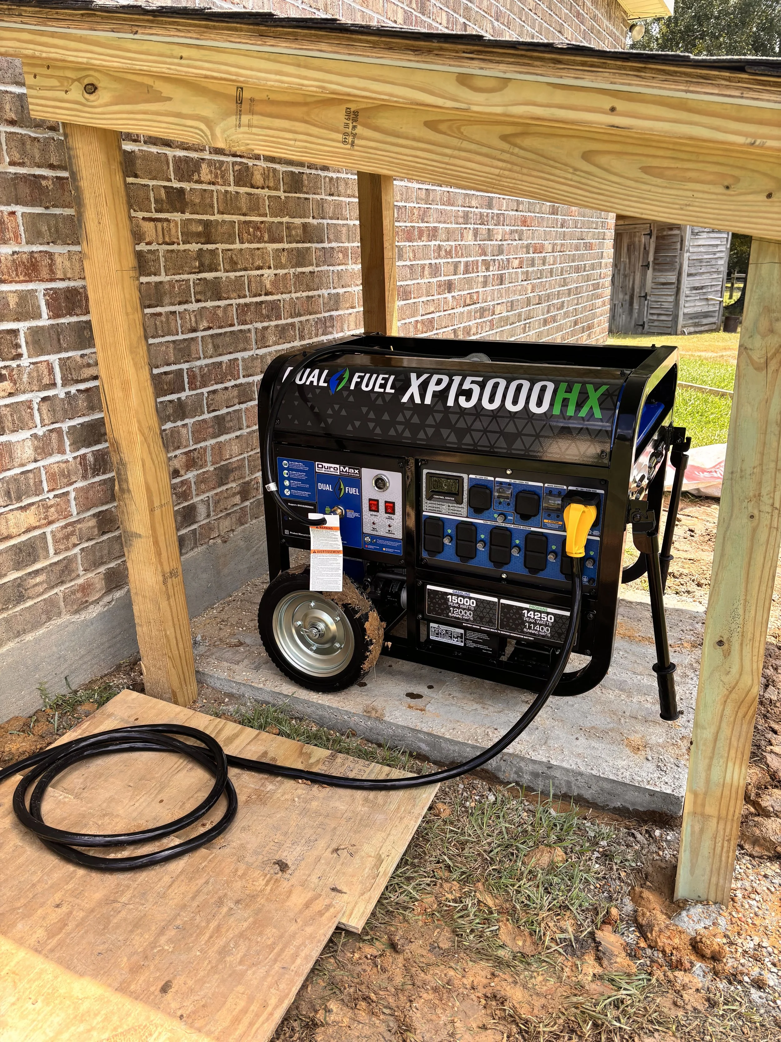 Portable generator outside a house under a wooden frame, with a brick wall behind it and a small patch of grass and dirt in the foreground.