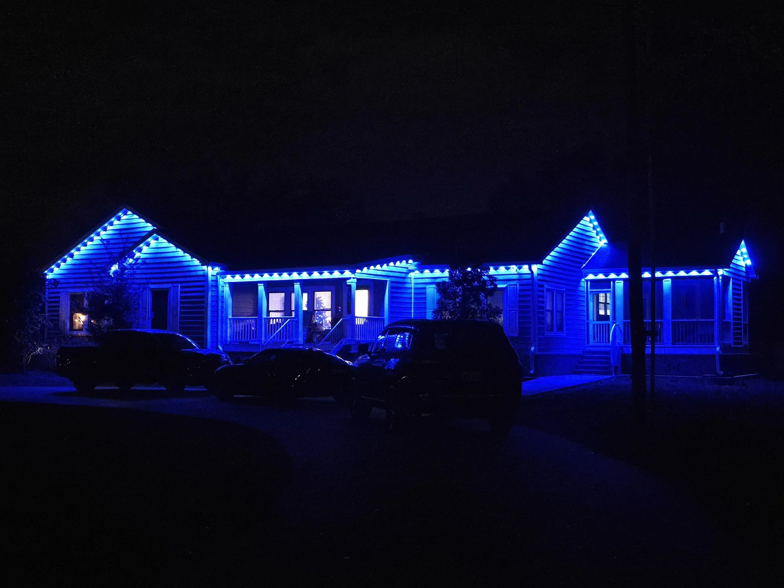 A house illuminated with blue holiday lights at night, with multiple cars parked in front.