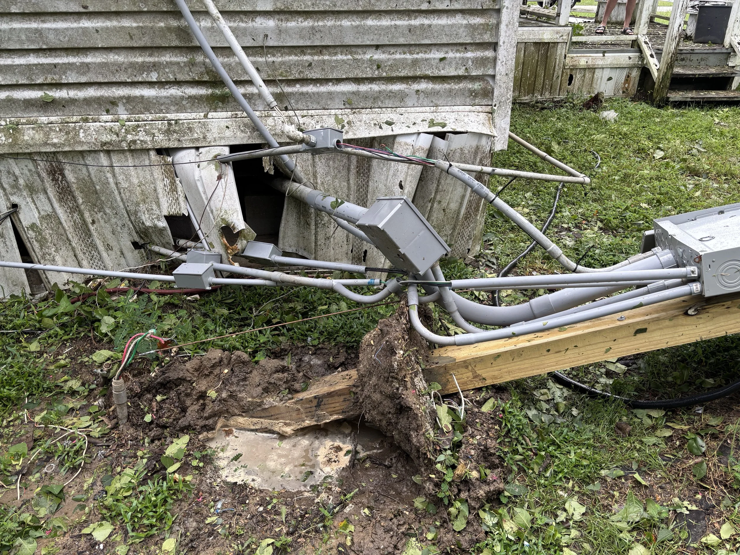 Electrical wires and conduit hanging loose outside a house with damaged siding, surrounded by grass and dirt, with exposed roots and a partial wooden support.