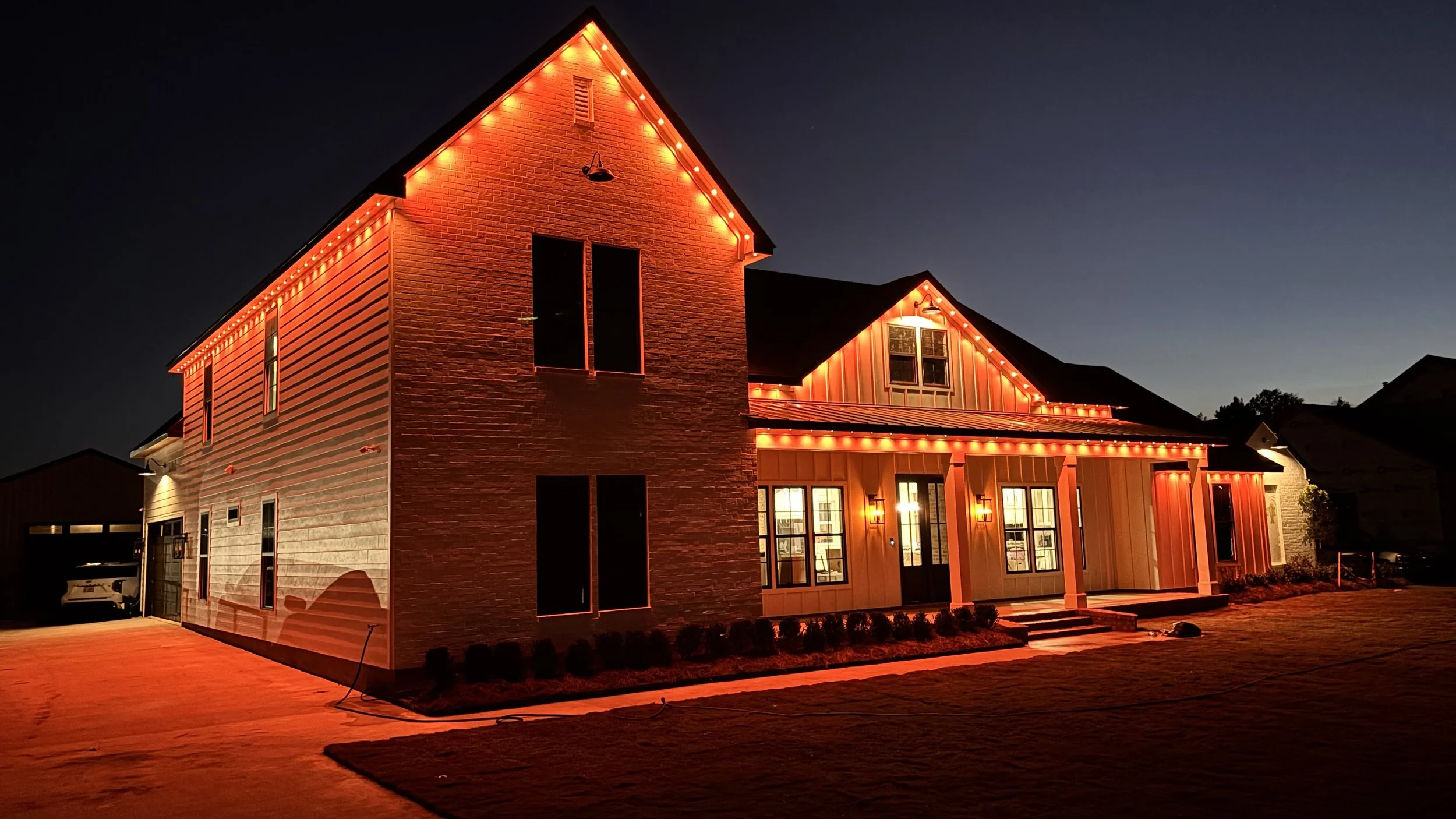 A large two-story house decorated with orange string lights at night, with illuminated windows and a driveway with a parked car.