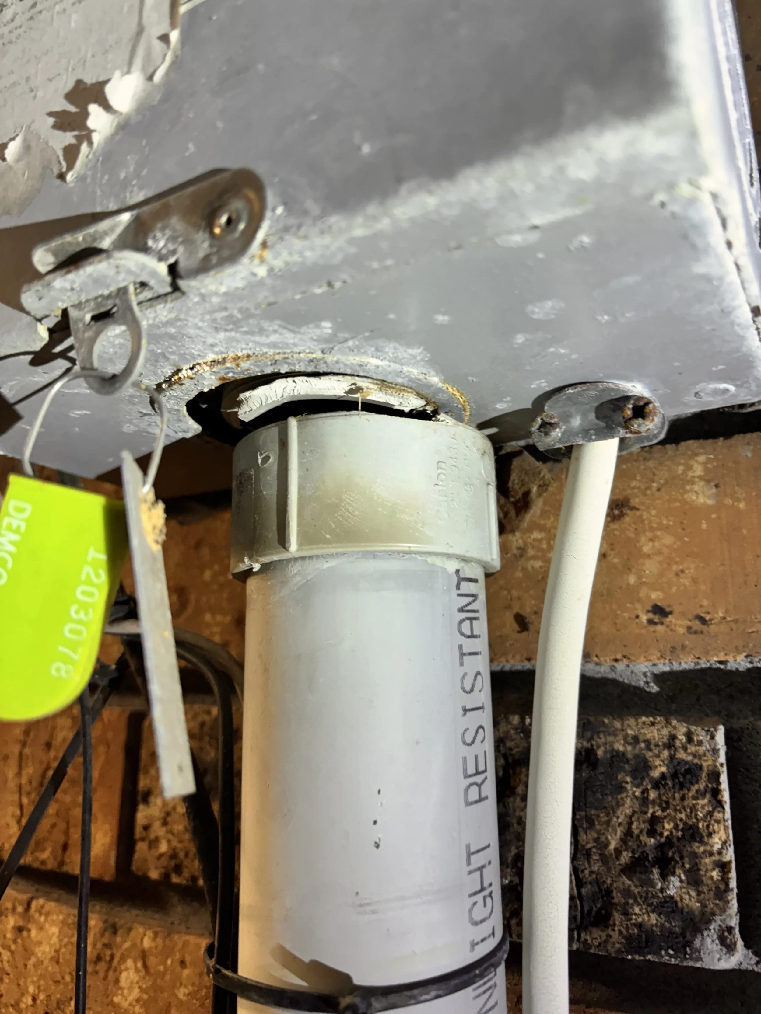 Close-up view of a plumbing fixture with a grey metal box, a white pipe labeled 'HIGH LIGHT RESISTANT,' and a white cable, mounted on a brick wall.