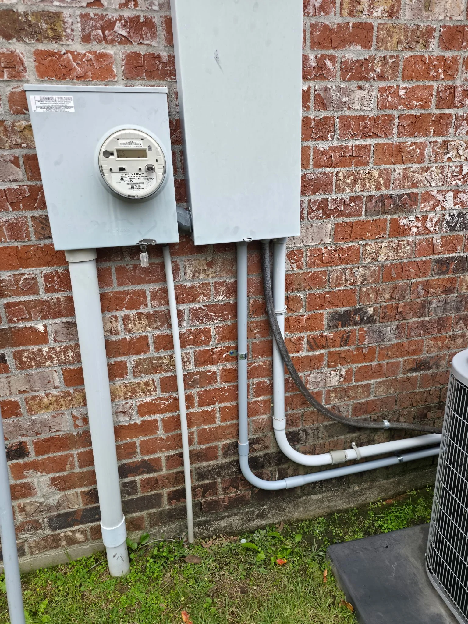 Outdoor view of an electric meter and electrical boxes mounted on a brick wall with conduit pipes leading to an air conditioning unit on a grassy ground.