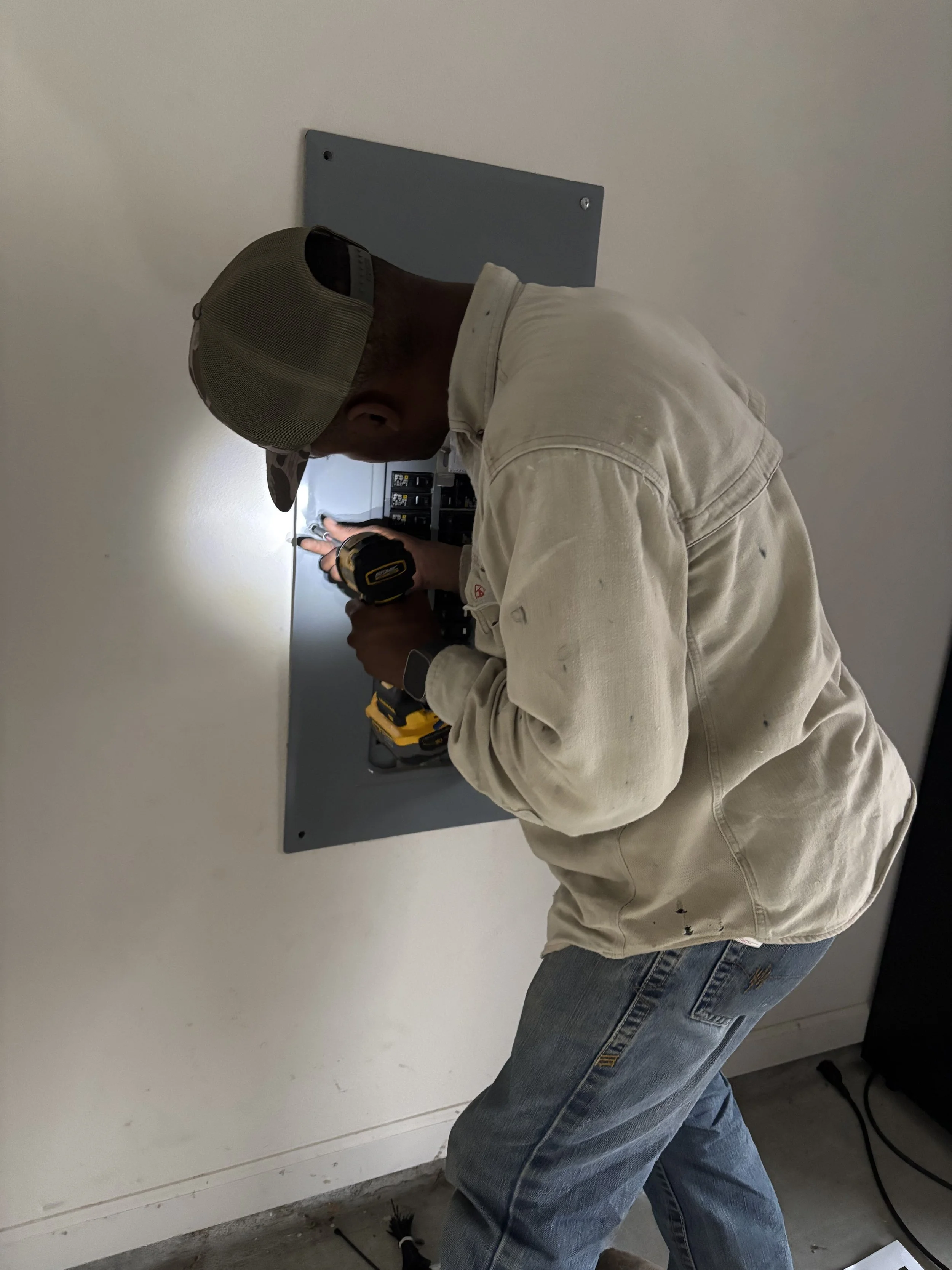 A man wearing a beige cap and work clothes installing or repairing electrical wiring inside an electrical panel on a white wall.