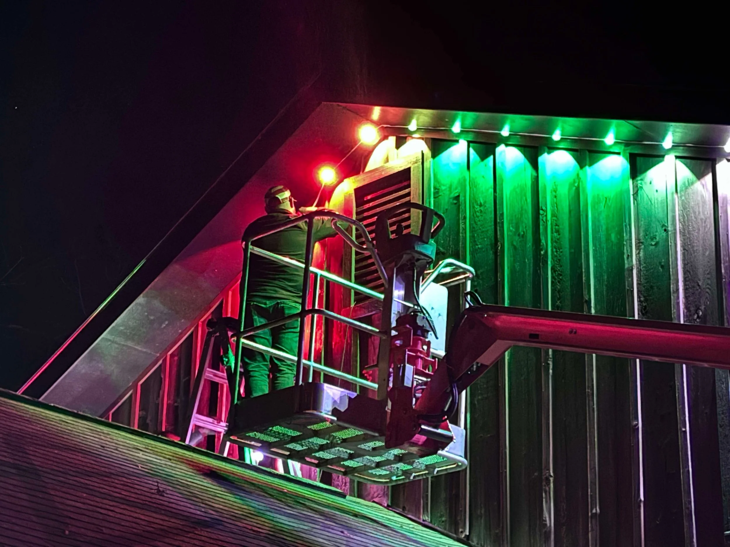 A worker on a cherry picker installs colorful holiday lights on a building at night.