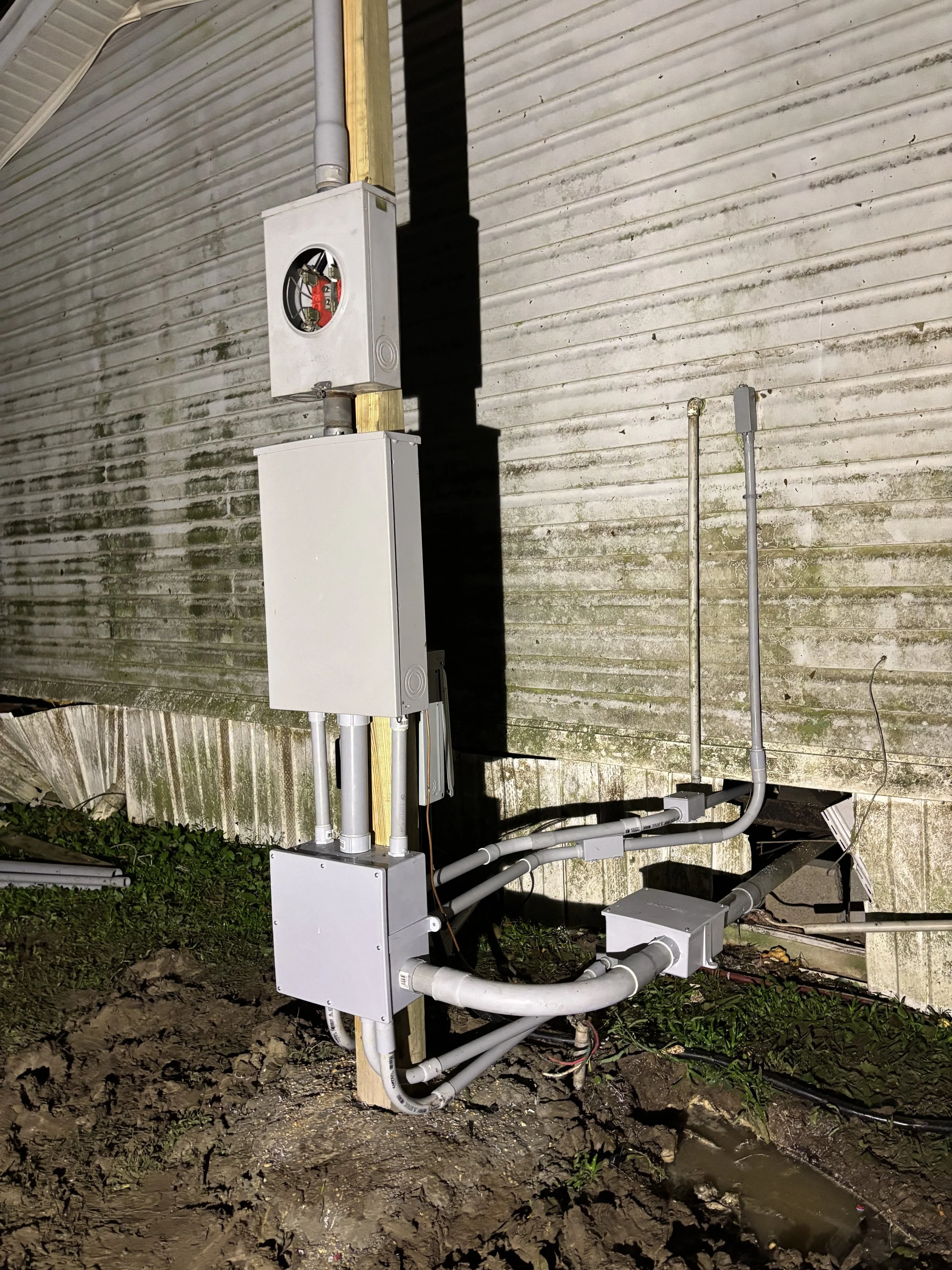 Electrical meter and conduit pipes installed next to a weathered wall with dirt and small plants at the base.