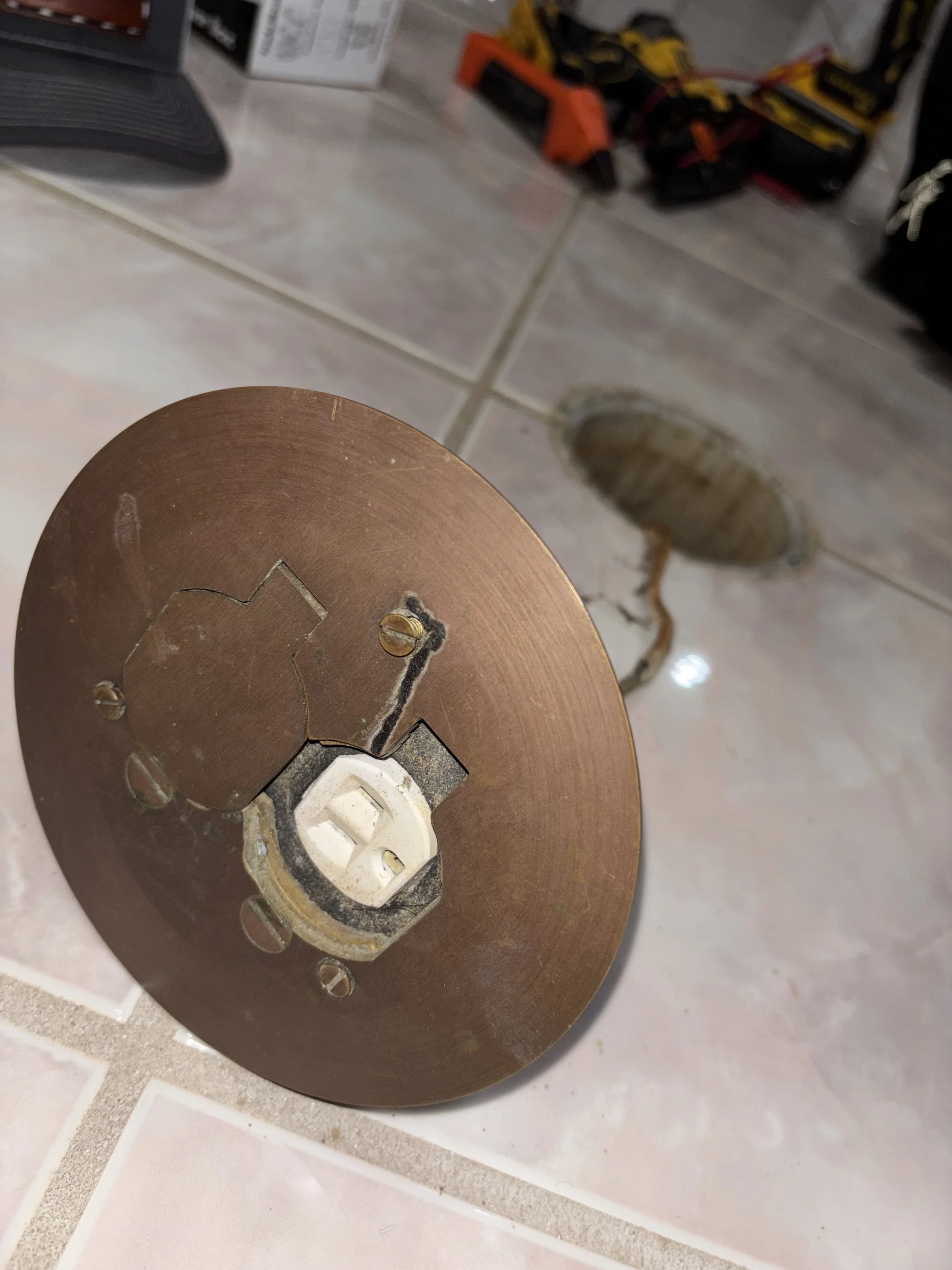 The image shows a disassembled circular power socket with a metal faceplate placed on a tiled floor, with tools and supplies visible in the background.