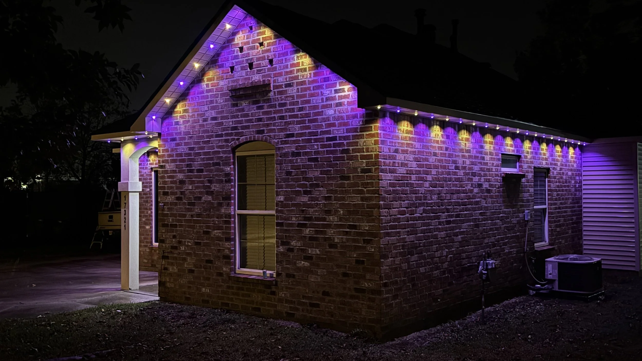 Nighttime photo of a brick house decorated with purple and yellow Christmas lights along the roofline and on the porch, with visible windows and an air conditioning unit outside.