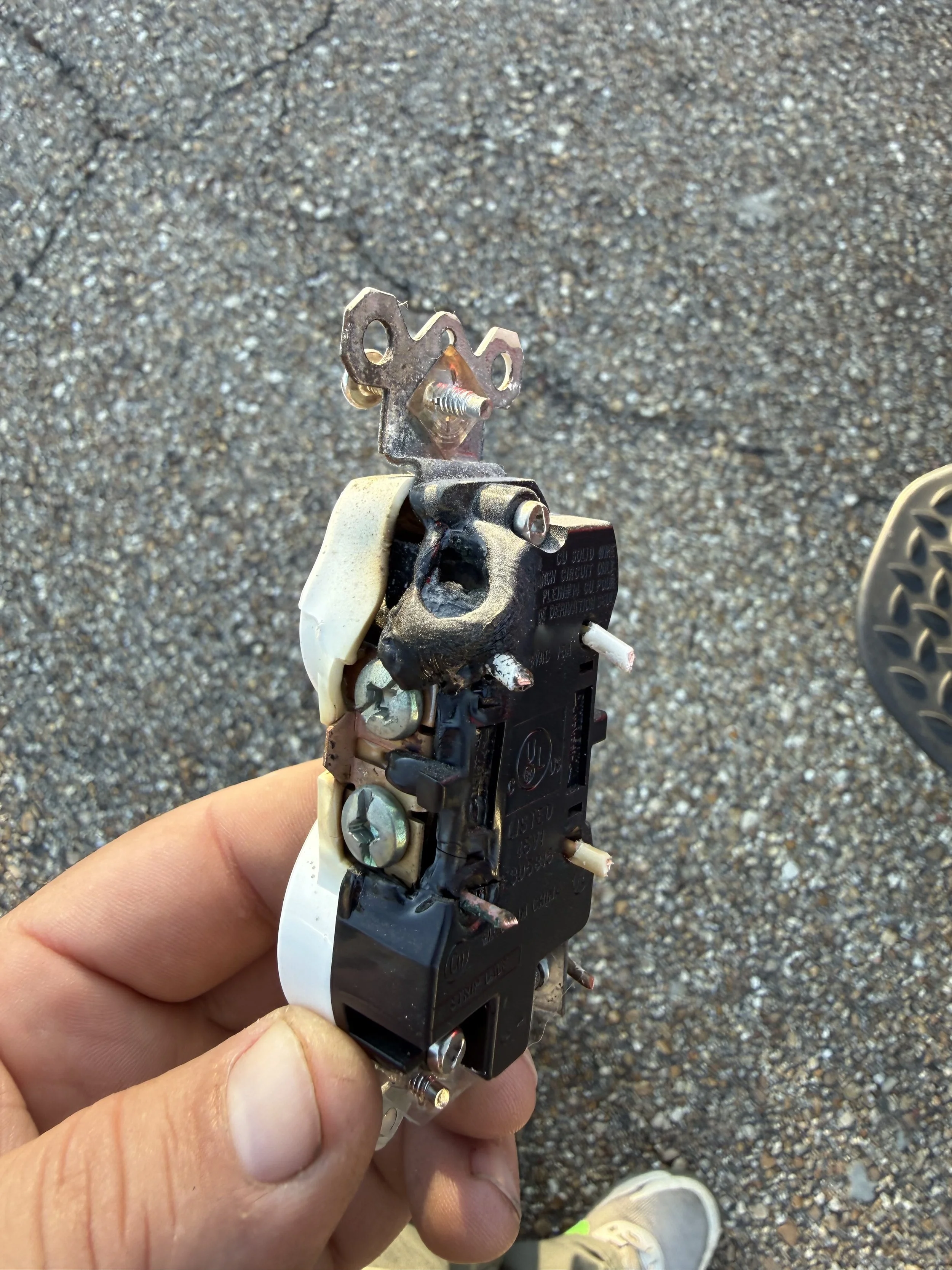 Close-up of a person's hand holding a black and silver electrical switch or relay with some burnt or damaged parts, mounted on a metal surface, with a gravel ground in the background.