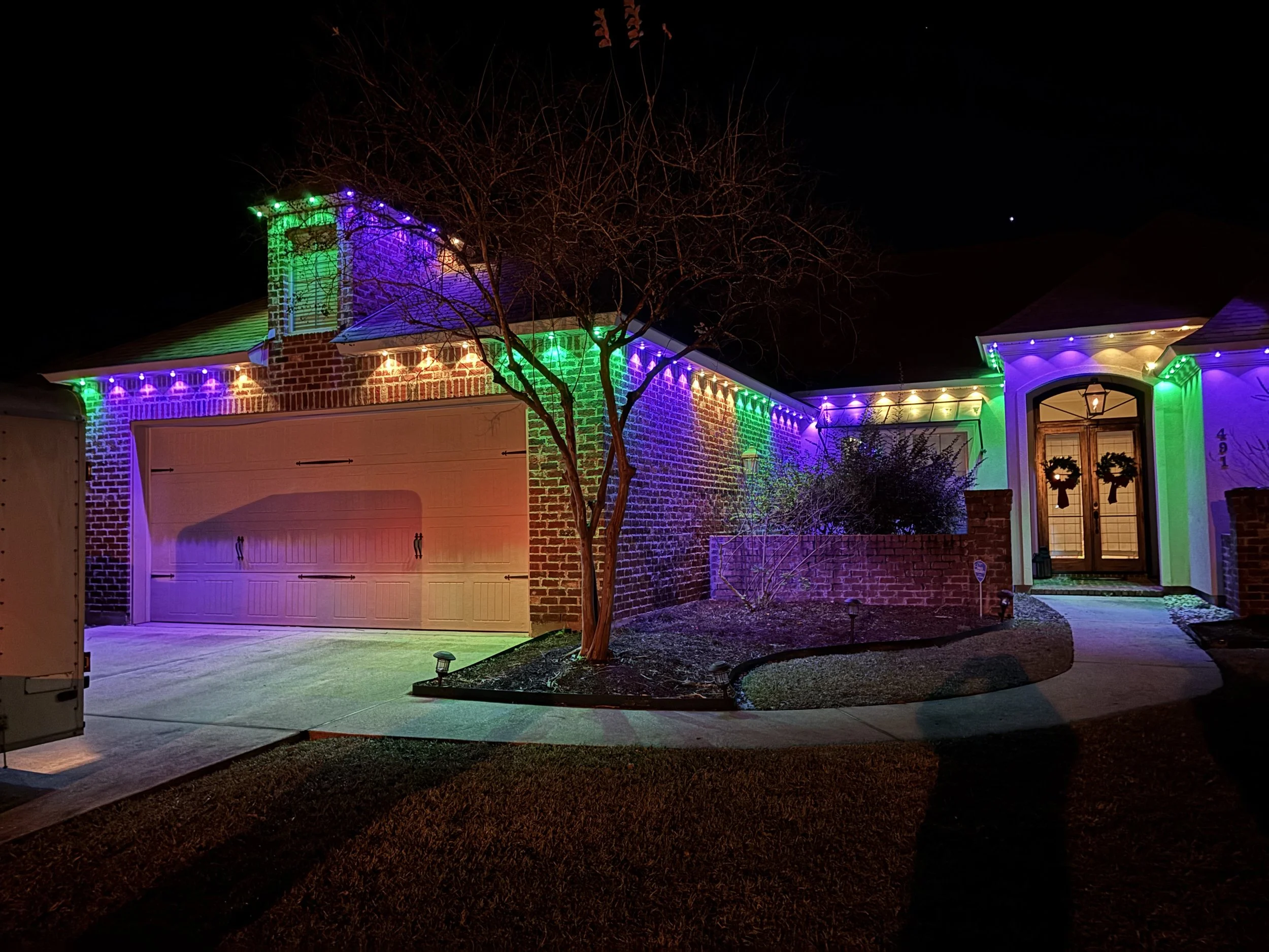 A house decorated with colorful holiday lights at night, featuring orange, purple, green, and yellow illumination on the roofline and surrounding the entrance.