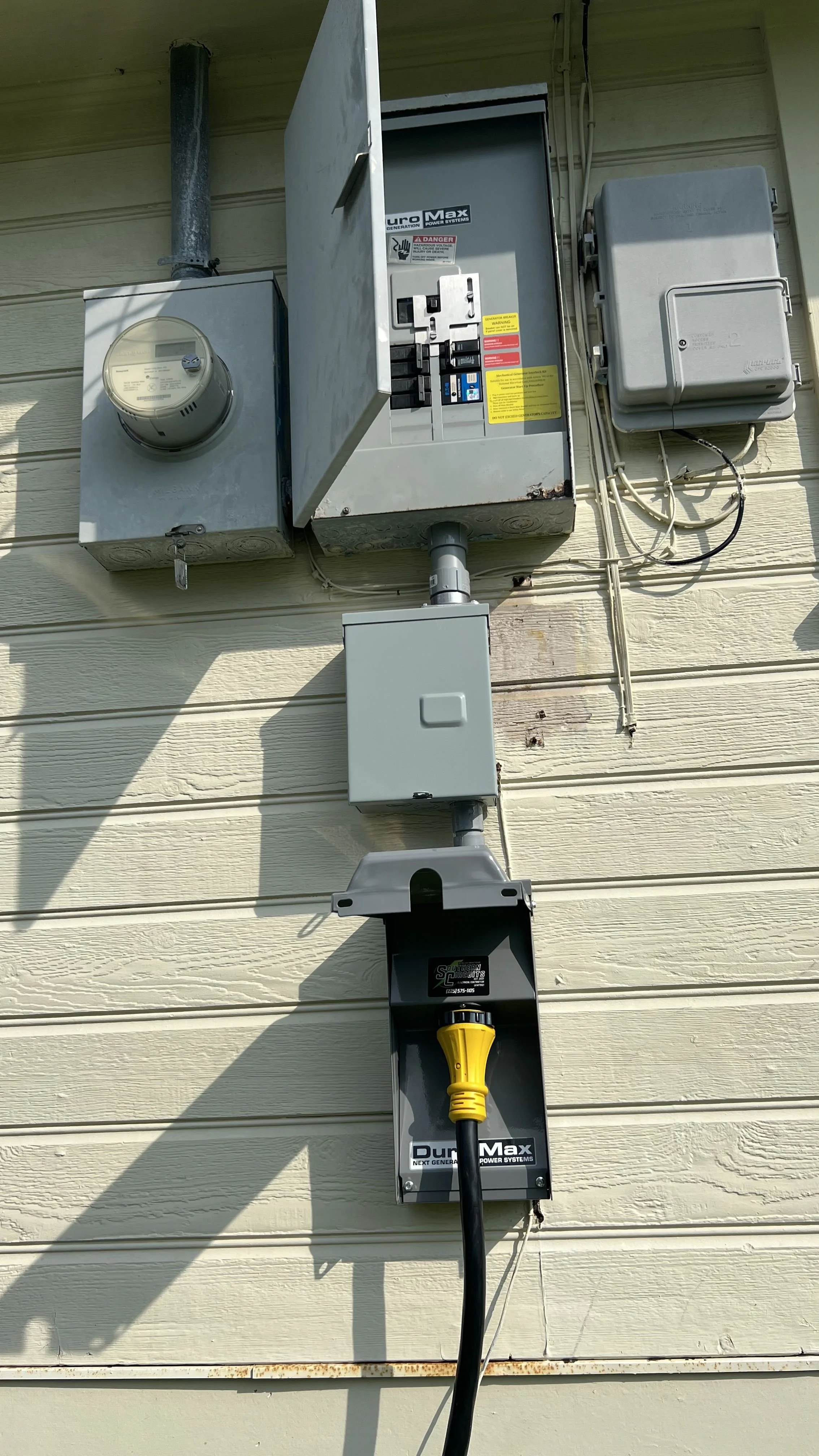 Electrical meter and control panels mounted on the exterior of a house with a power cord plugged into a power outlet.