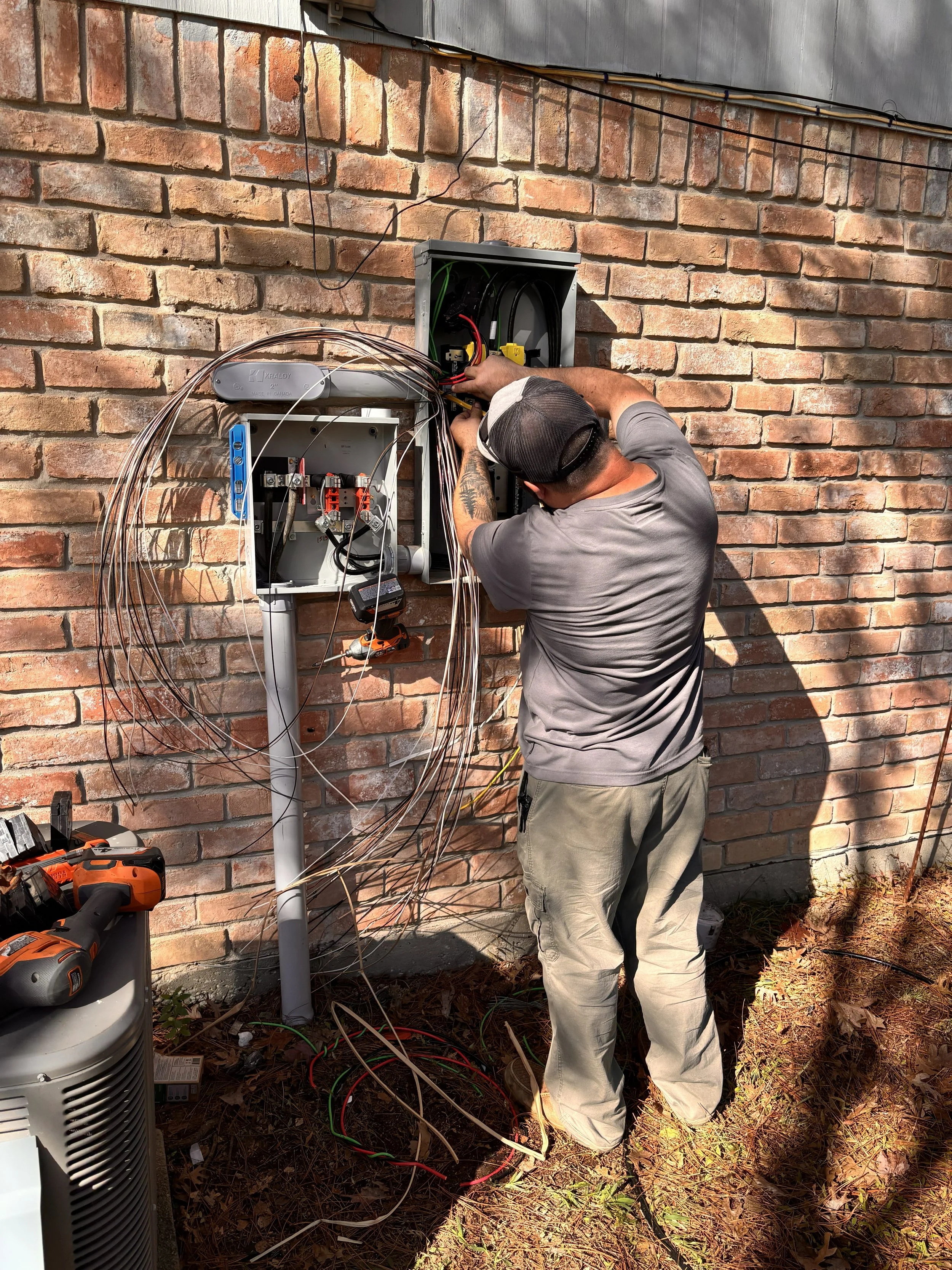 A technician working on outdoor electrical wiring inside an open electrical panel mounted on a brick wall