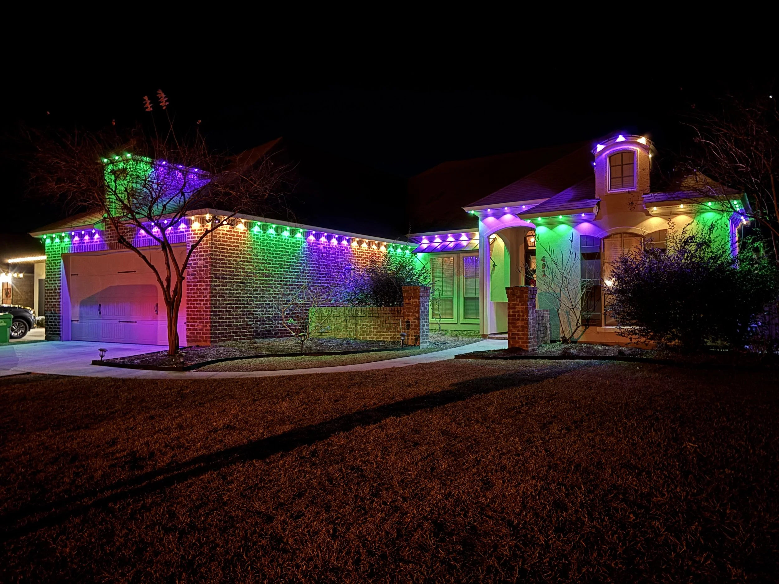 A house decorated with colorful Christmas lights at night, including green, purple, and blue lights outlining the roof and walls, with a leafless tree in the front yard.